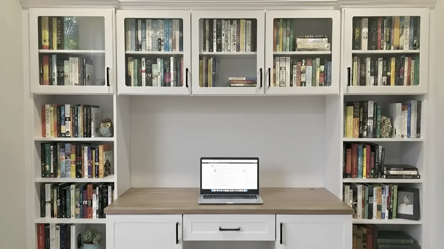 A white bookshelf with glass doors filled with various books. A laptop is open on a wooden desk in front of the bookshelf.