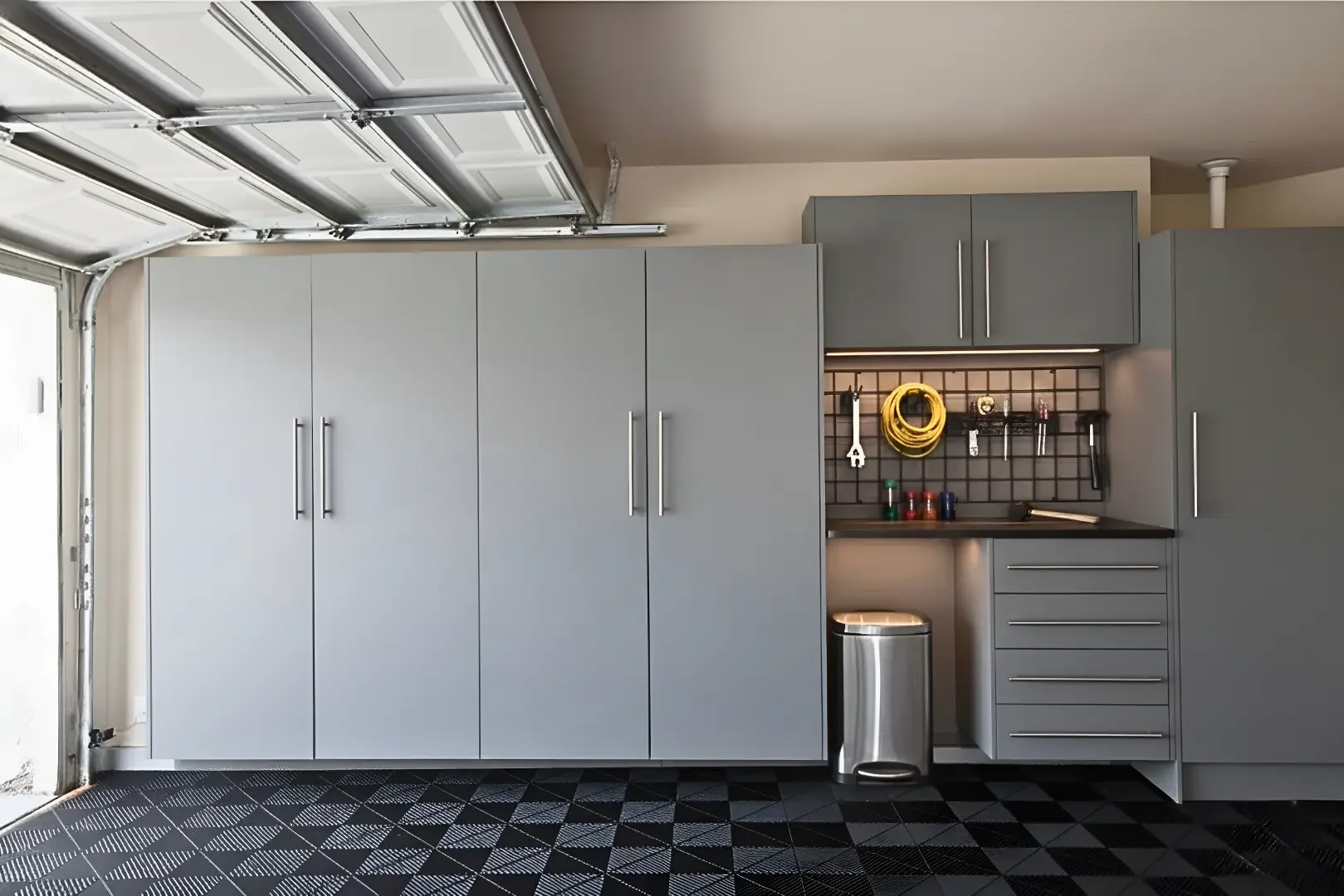 A garage with gray cabinets, a pegboard with tools and supplies, a stainless steel trash can, and patterned black flooring.