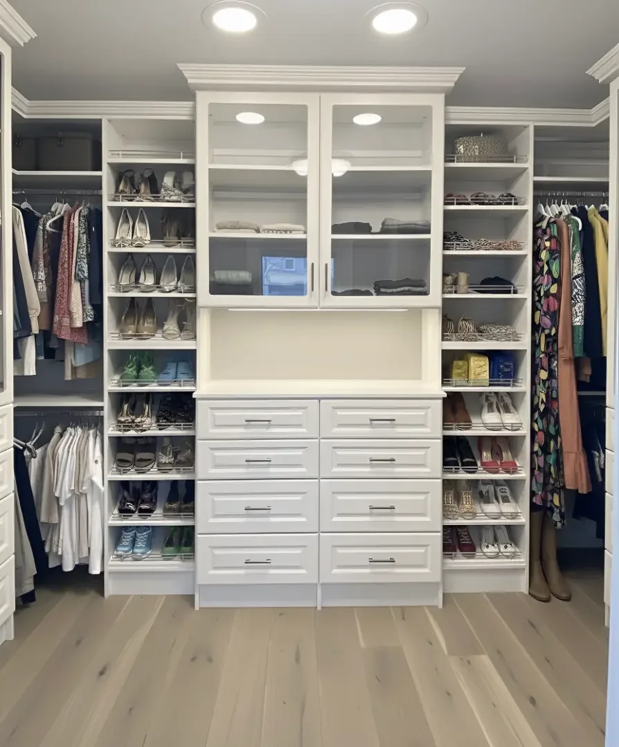 A walk-in closet with white cabinetry, glass-front cabinets in the center, and open shelves on each side filled with shoes, handbags, and clothing.