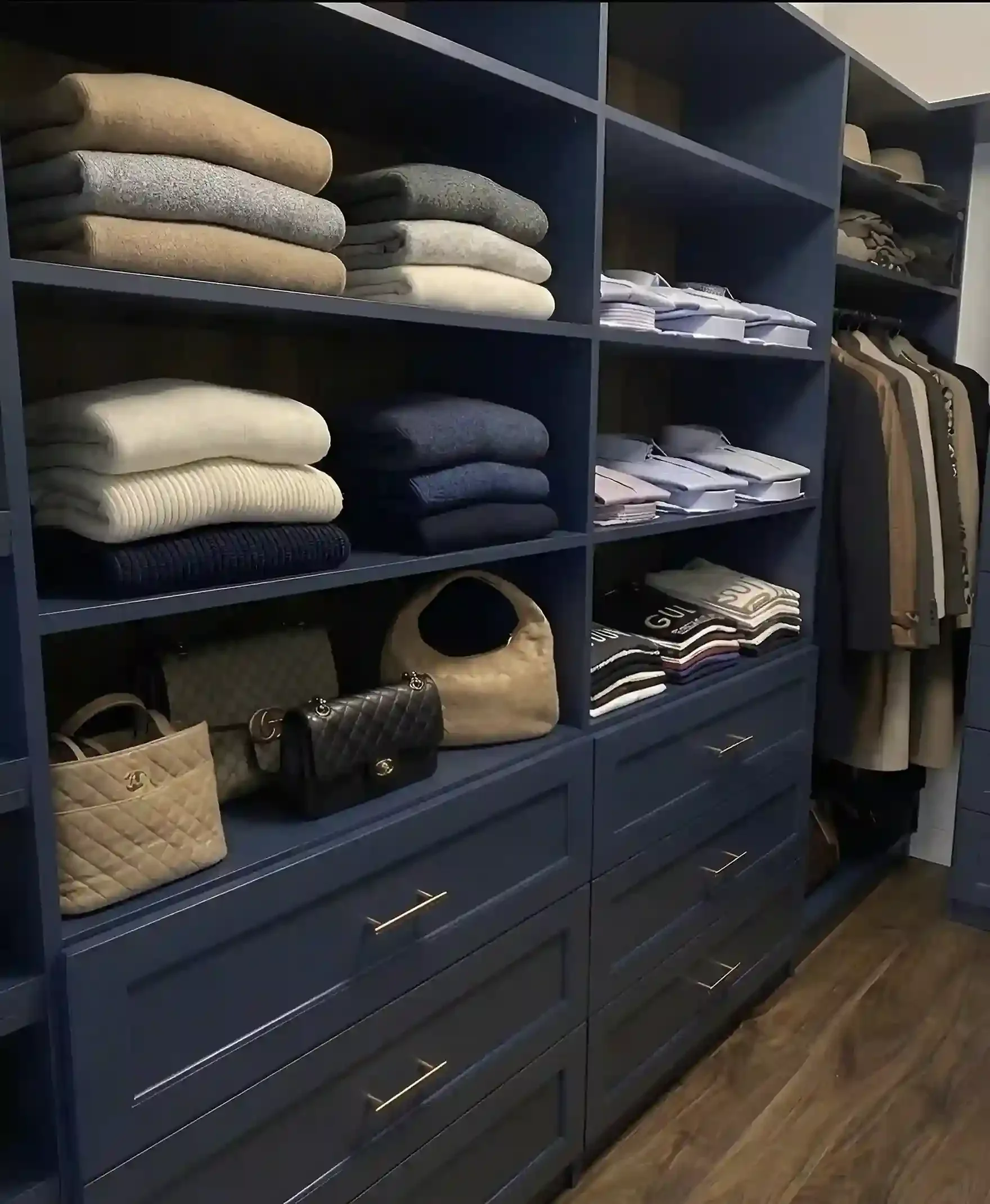 A closet with shelves containing neatly folded sweaters and stack of clothes, with handbags and shoes on the lower shelves.