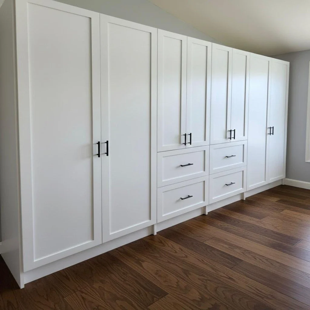 A row of white built-in cabinets with black handles in a room with hardwood floors.