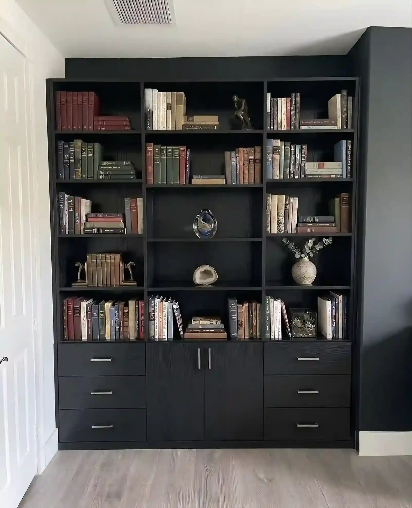 A black bookshelf filled with books and decorative items stands against a grey wall in a room with a wooden floor.