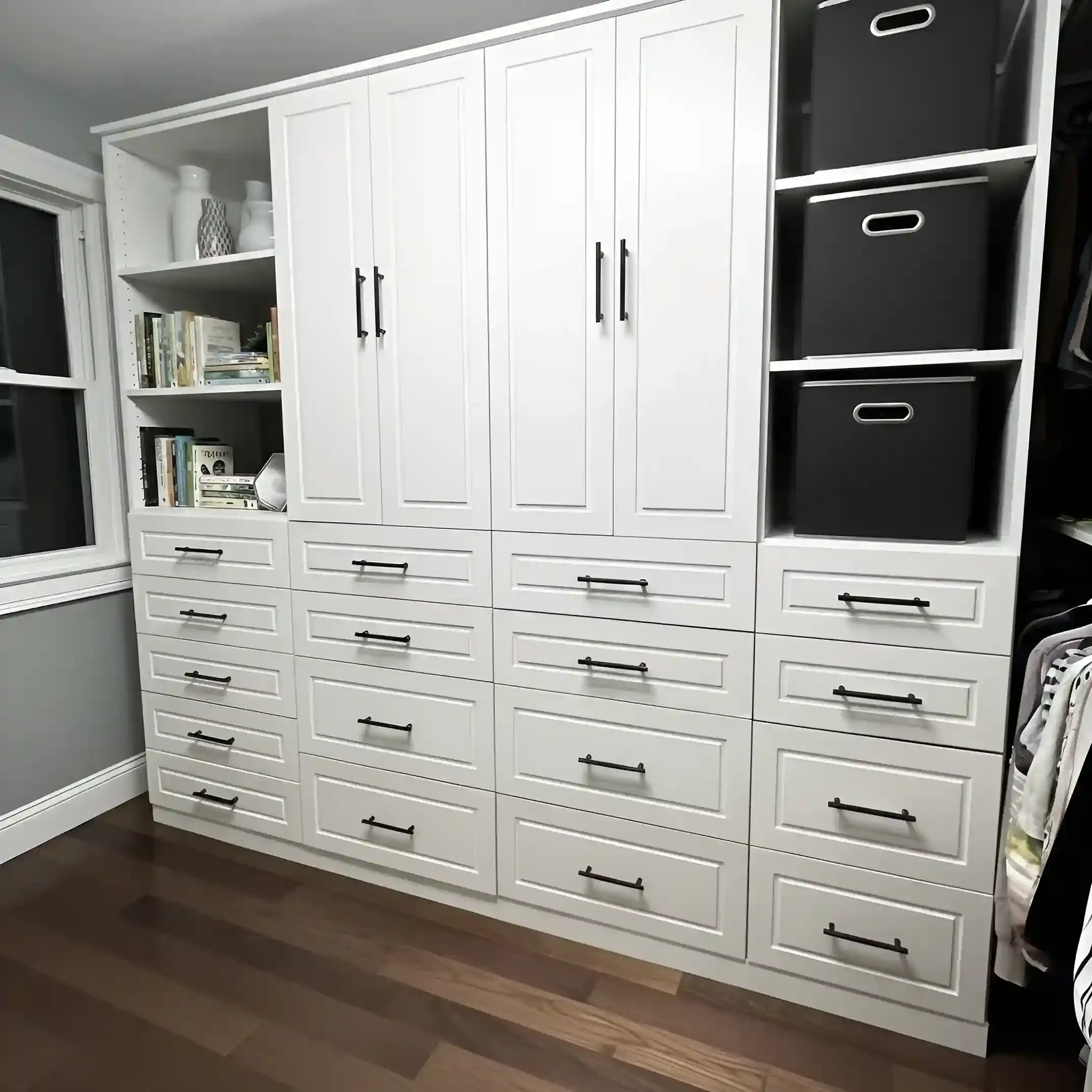 White wardrobe with multiple drawers and cabinets, black handles, and open shelves containing a few books and decorative items, situated in a room with wood flooring and a window.