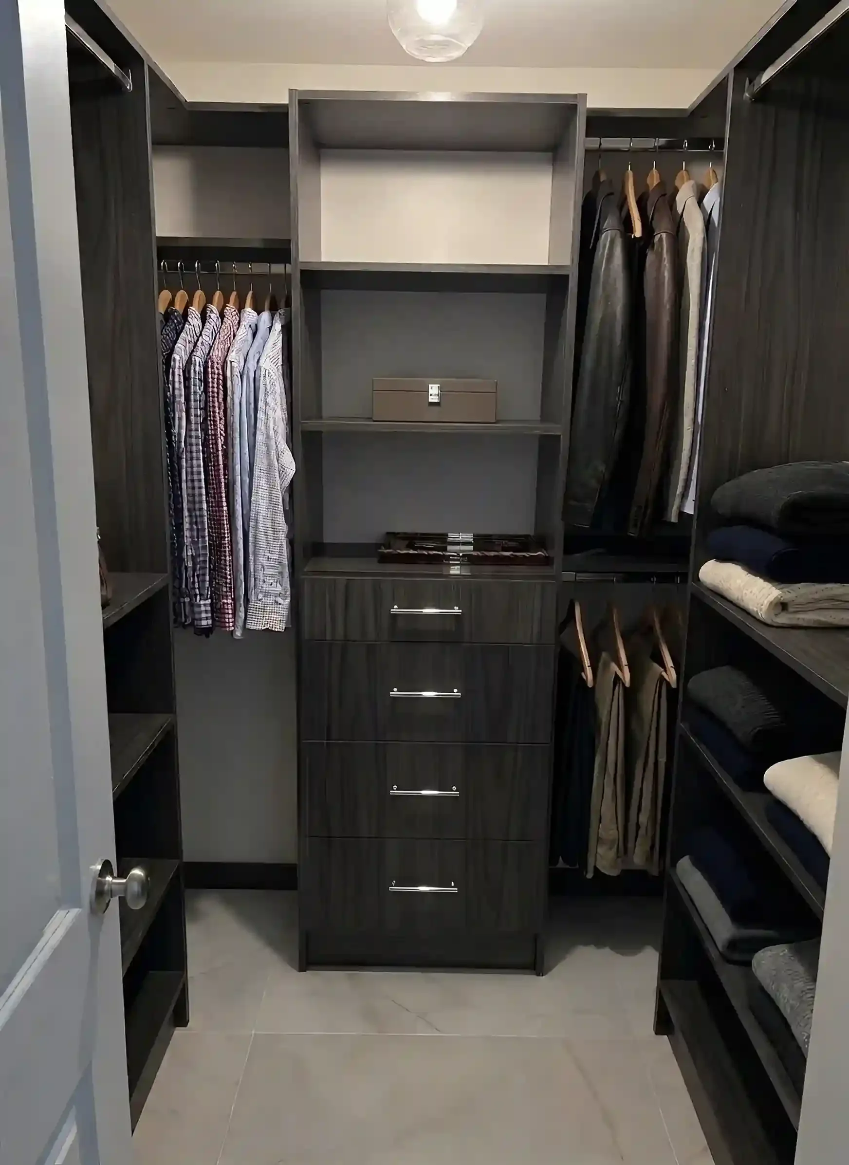 A walk-in closet with black wooden shelves and hanging clothes, including shirts, jackets, and folded towels, with a gray tiled floor and a ceiling light fixture.
