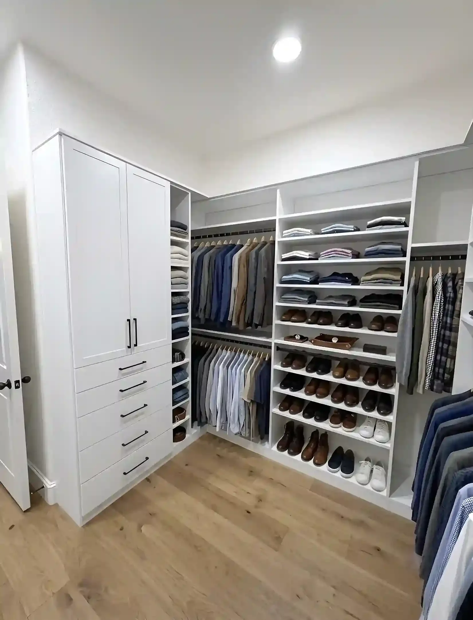 A walk-in closet with white shelving units filled with folded clothes, hanging shirts and pants, and shoes lined on shelves and the floor. The room has wooden flooring and bright lighting.