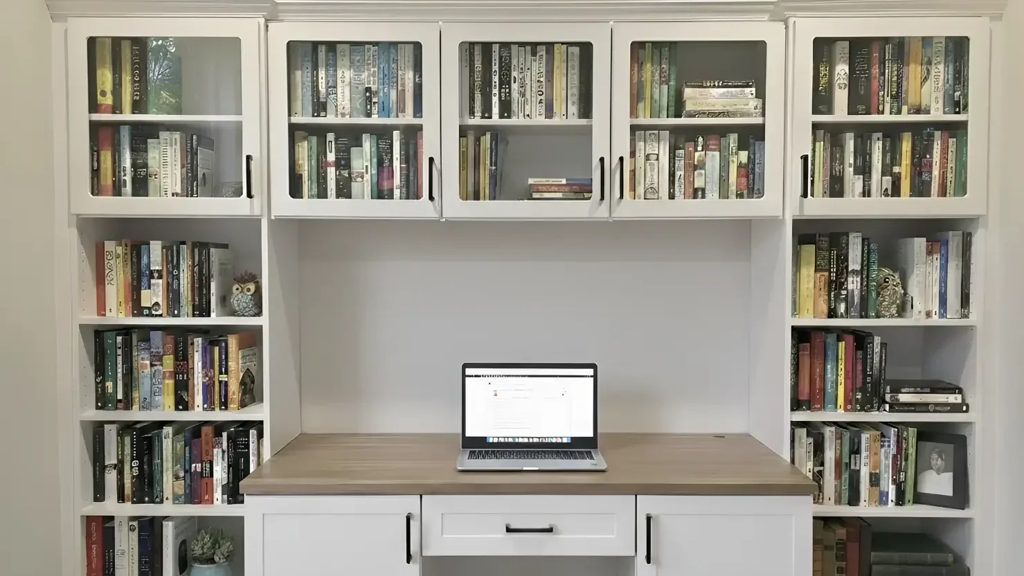 White bookshelf with glass doors filled with books, a wooden desk with a laptop in the center, and small decorative owl and skull figurines on the bookshelves.