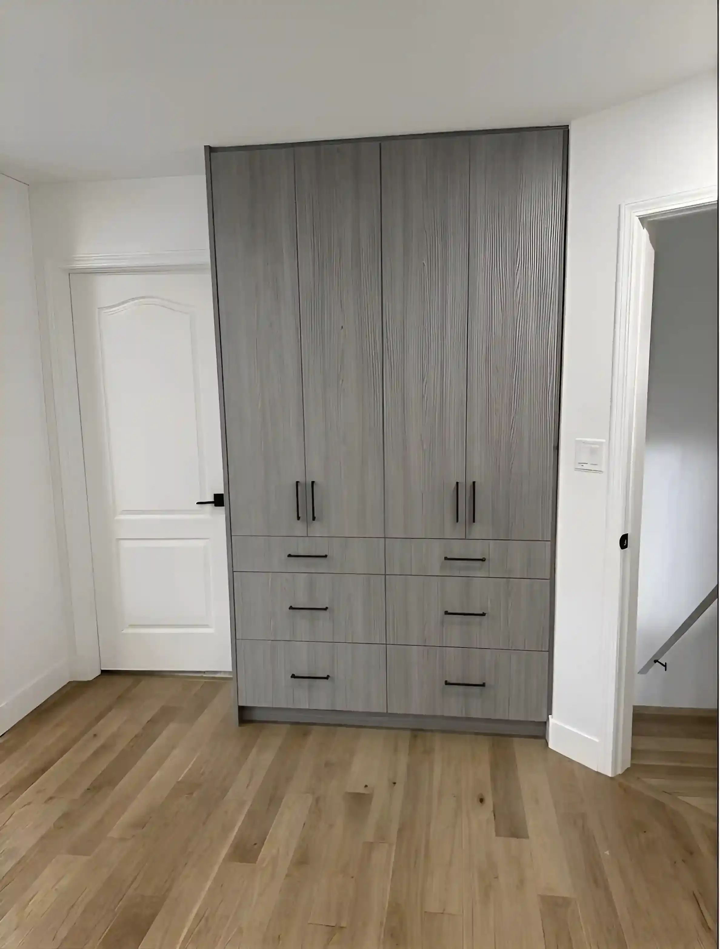A tall, modern kitchen cabinet with a wood grain finish and black handles, located against white walls with crown molding, and a wooden floor.