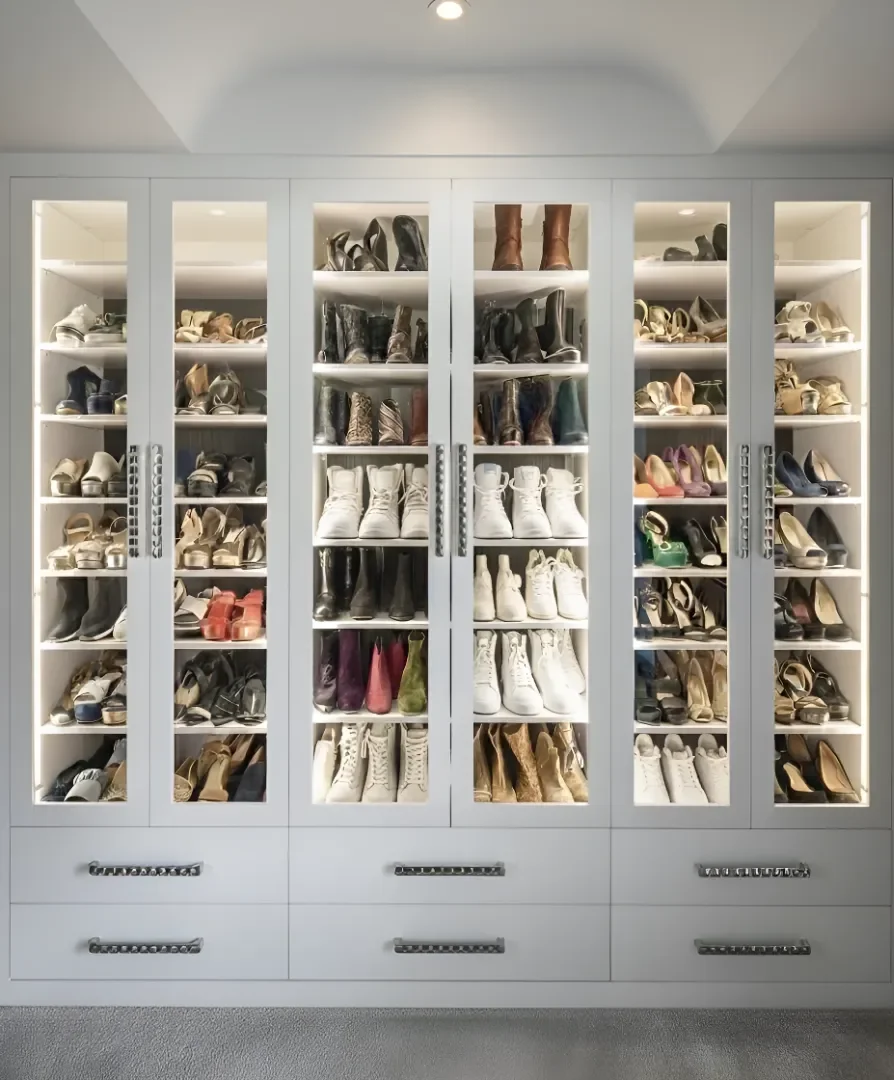 A white shoe cabinet with glass doors filled with various men's and women's shoes, including sneakers, boots, sandals, and high heels, neatly organized on shelves.