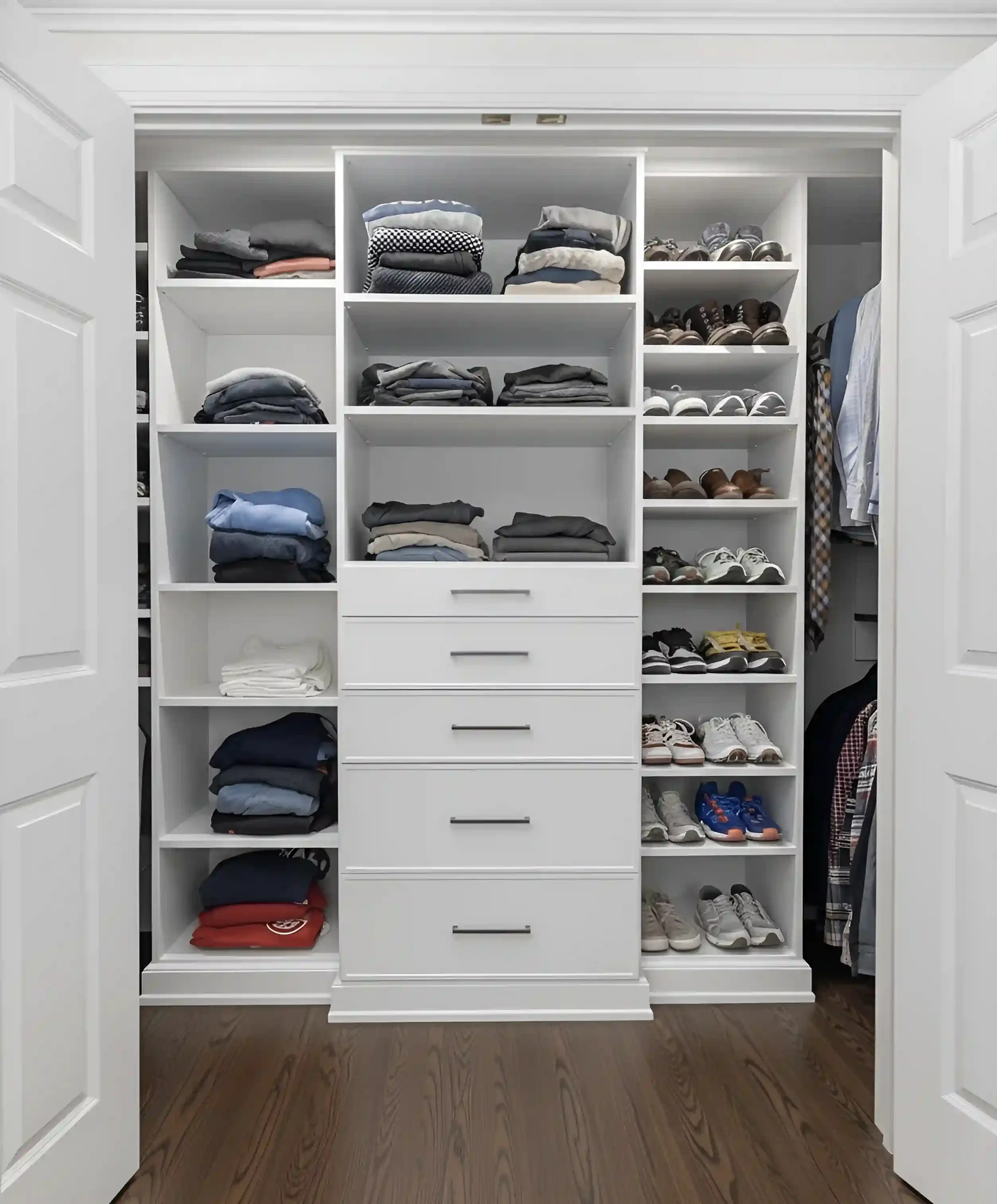 A neatly organized white walk-in closet with shelves filled with folded clothes on the left and right sides, and a central set of drawers. The floor is wooden, and the closet doors are open.