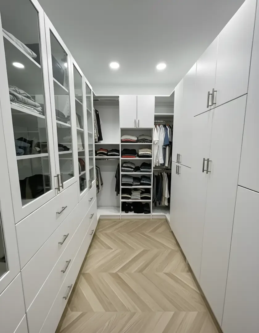 A walk-in closet with white cabinets, glass-front drawers, and open shelves filled with folded clothes and hanging garments, with a chevron-patterned wooden floor and recessed ceiling lights.