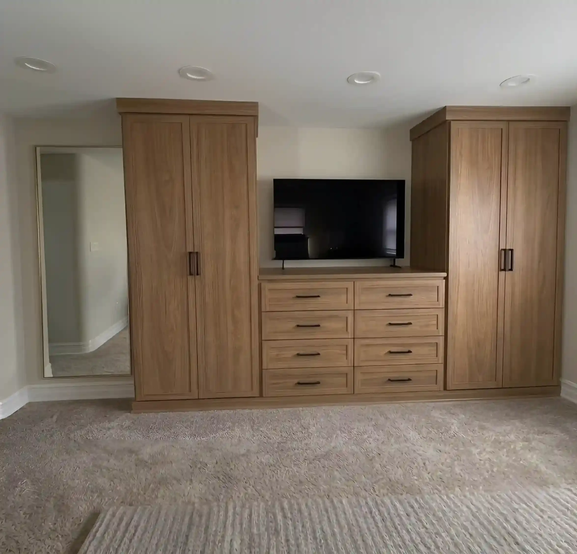 A room with beige carpeted flooring and a wooden wall unit with a TV in the middle. There is a large mirror on the left wall and a small doorway in the back left corner.