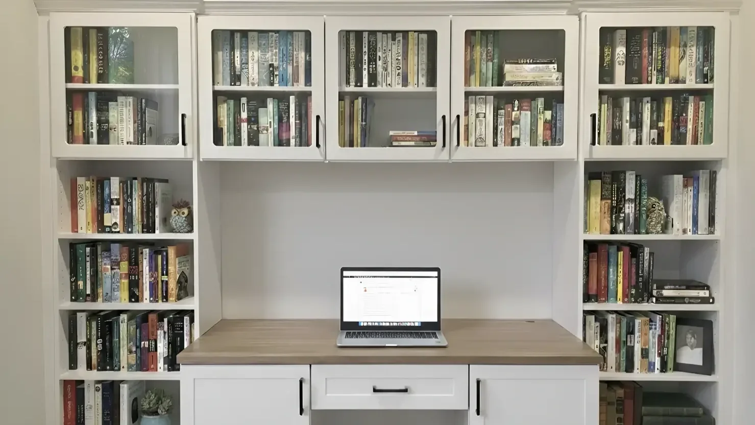 A white wooden bookshelf with glass doors filled with books and decorative items, a wooden desk with a laptop on it, and a cabinet with a drawer underneath.