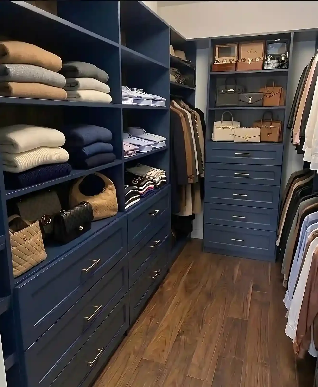 A walk-in closet with navy blue shelves and drawers holding folded clothes, handbags, and boxes, with hanging shirts and trousers on the right side.