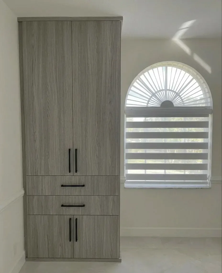 A tall, modern grey wood cabinet with black handles next to a window with a half-circle fan-shaped window covering and horizontal blinds, in a light-colored room.