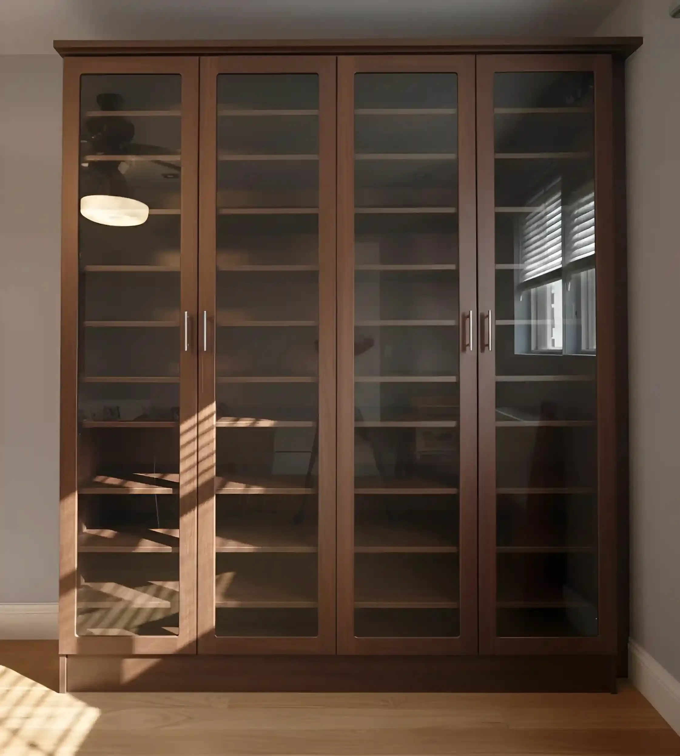 A large wooden display cabinet with glass doors, empty shelves, and silver handles, positioned against a wall with sunlight and window reflections visible.