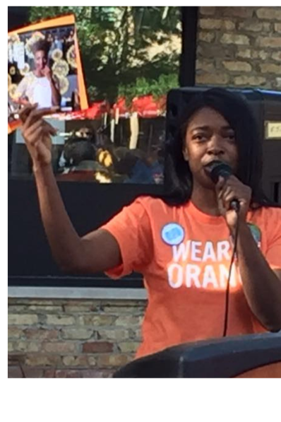 Woman speaking into a microphone while holding a framed photo of a person at an outdoor event.