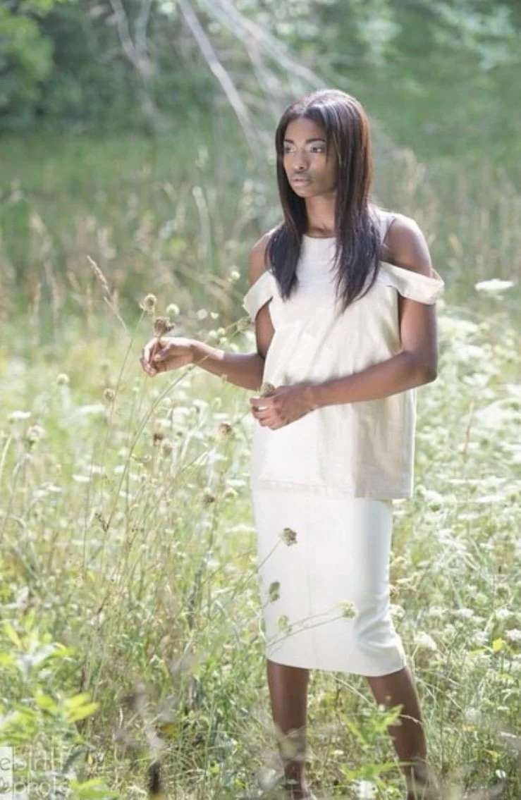 A young woman stands in a field of tall grass and wildflowers, holding a dandelion, with trees and greenery in the background.