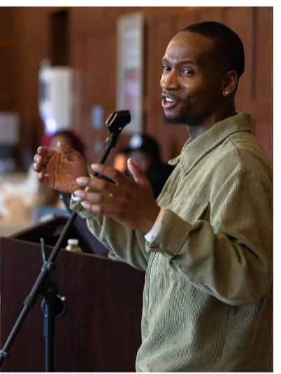 Man speaking at a microphone in a cozy indoor setting, possibly a restaurant or café, with wood-paneled walls.