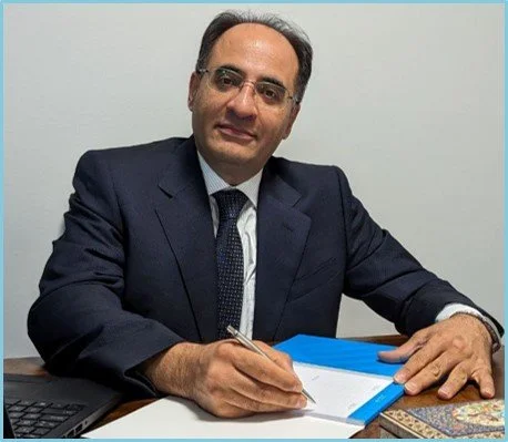 A man in a dark suit and glasses sitting at a desk with a pen, a closed blue folder, and a laptop.