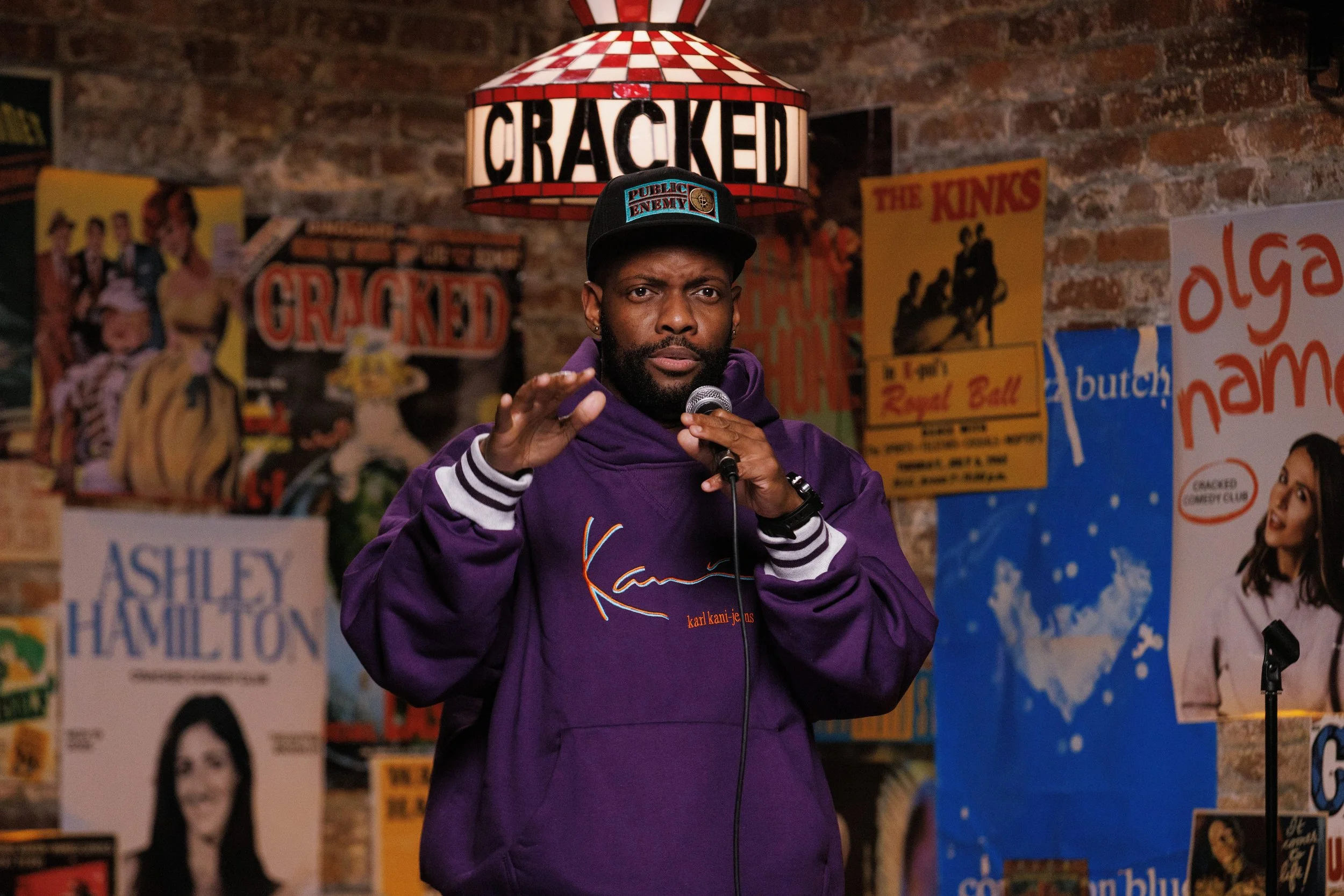 A man with a beard in a purple jacket is speaking into a microphone in a comedy club. Behind him, there are posters, including one that says 'CRACKED' and other theater posters on a brick wall.