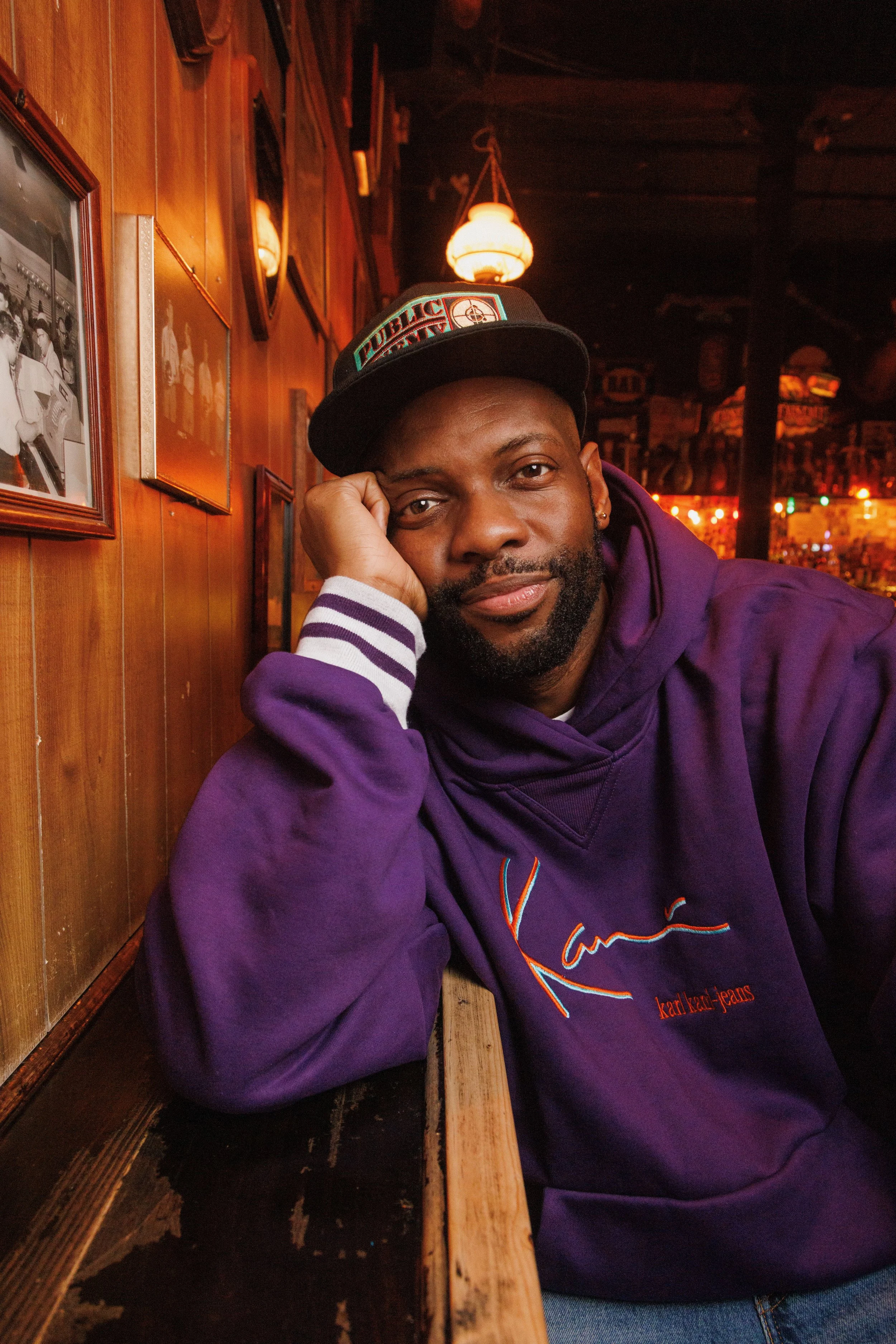 A man in a purple hoodie with 'Karl Kani' embroidered on it, wearing a black cap with colorful text, sitting in a cozy bar with wooden wall paneling and vintage photos, smiling and resting his head on his hand.