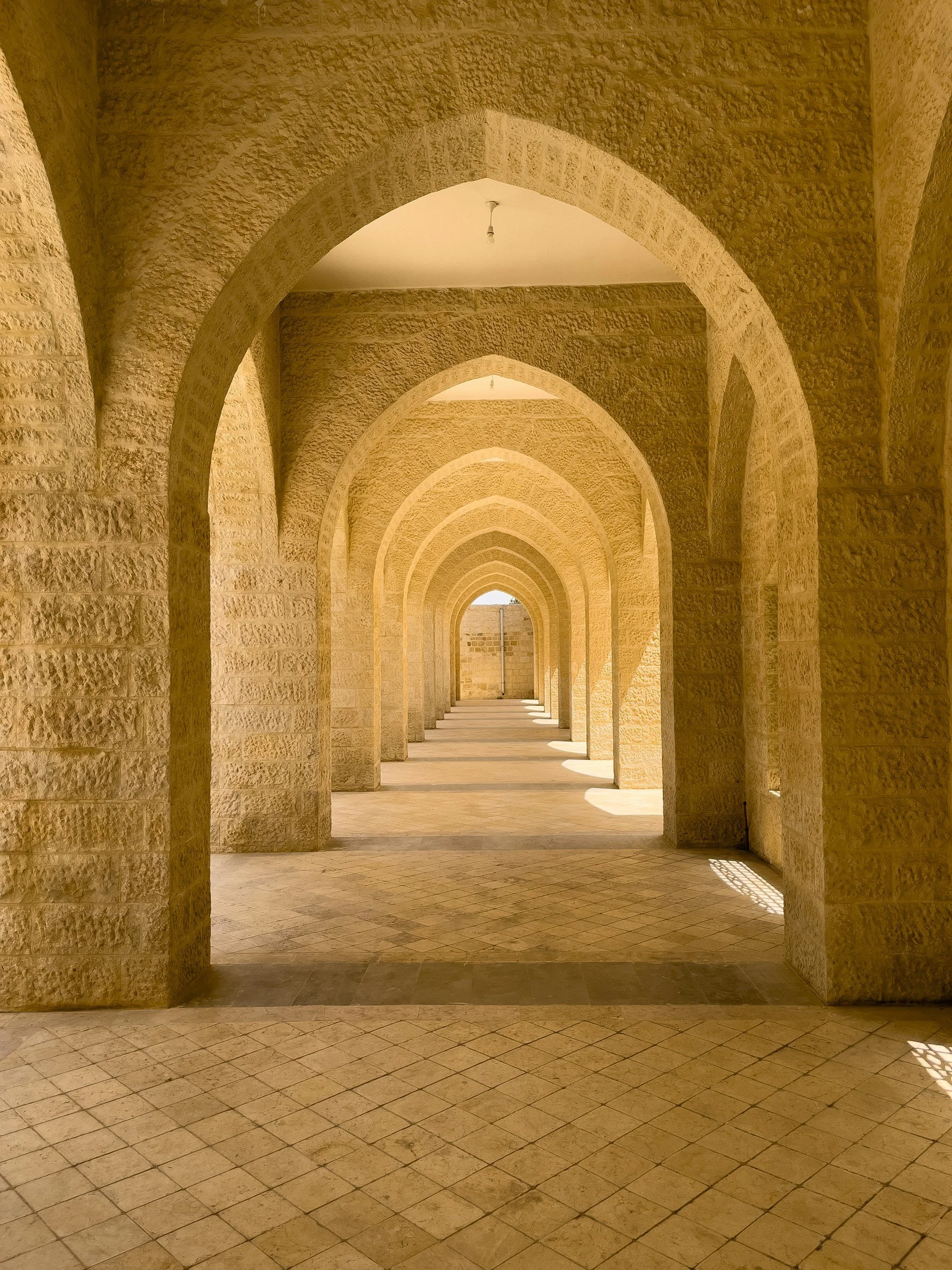 A corridor with repeating stone arches and tiled floor, sunlight casting shadows.