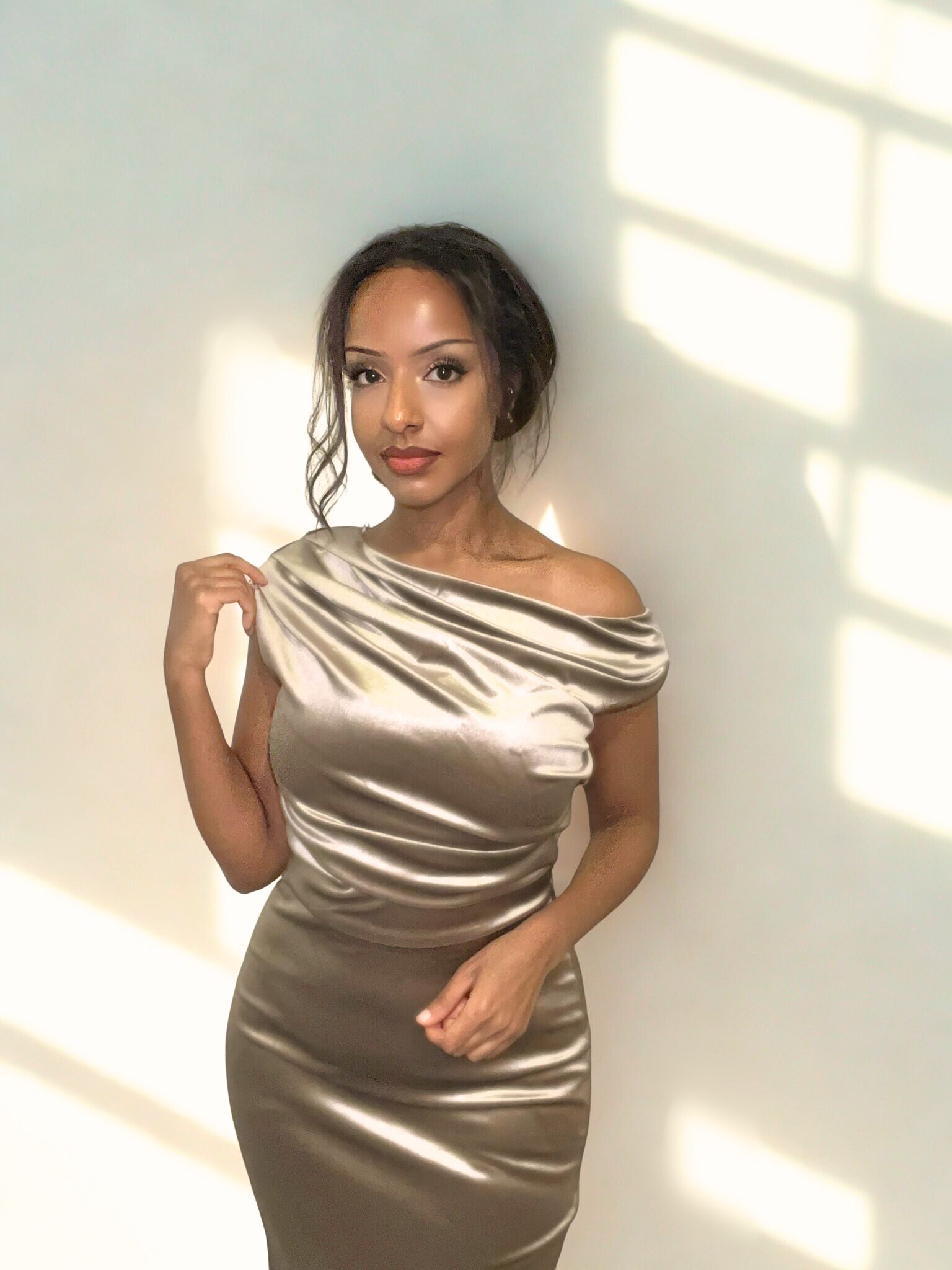 A woman with long dark hair in loose waves wearing a shiny, form-fitting, off-the-shoulder satin dress standing against a plain light-colored wall with sunlight and window shadows.