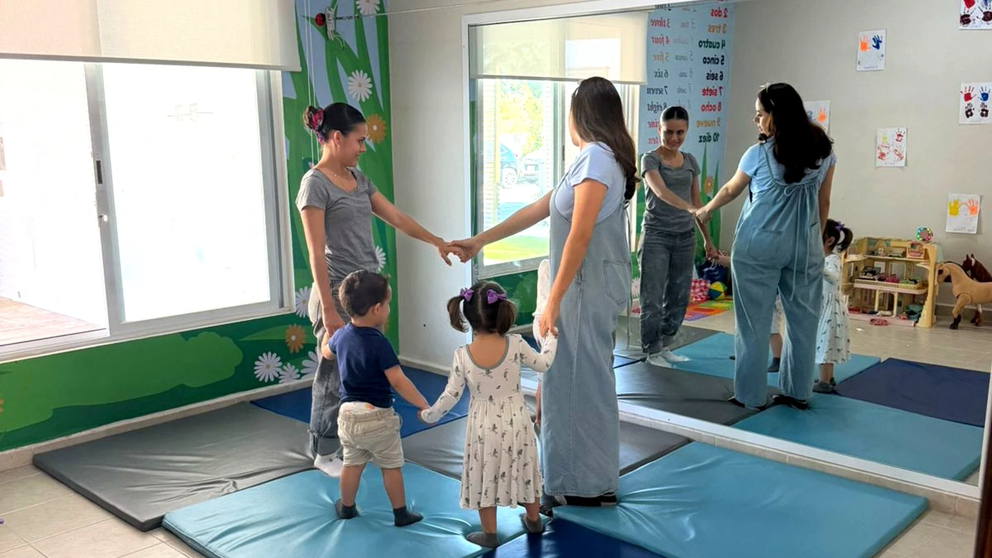 Niños y maestras en un ambiente de guardería o clase infantil, de pie sobre colchonetas, jugando mientras sostienen las manos en círculo.
