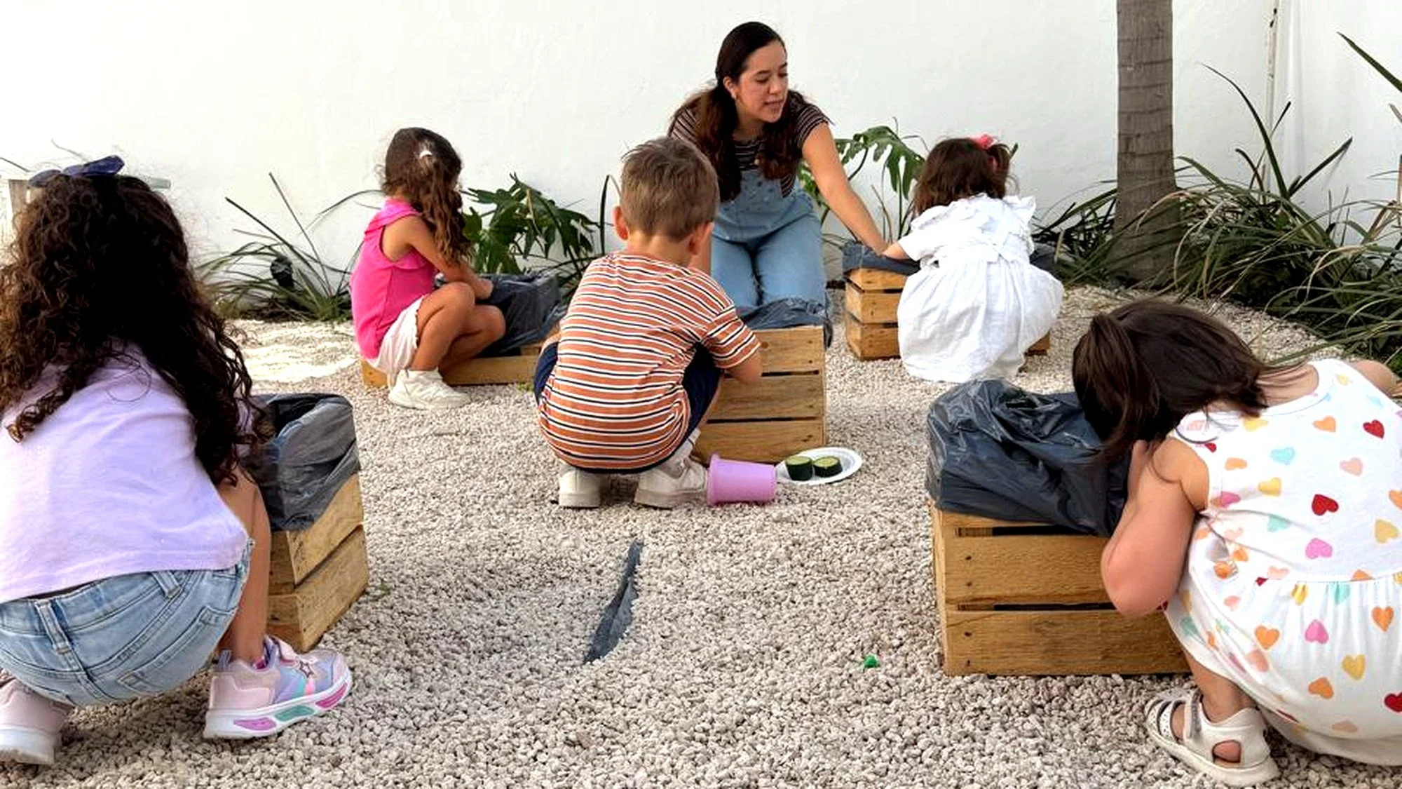 Niños y una mujer en un jardín, sembrando plantas en cajas de madera mientras están sentados en piedras blancas.