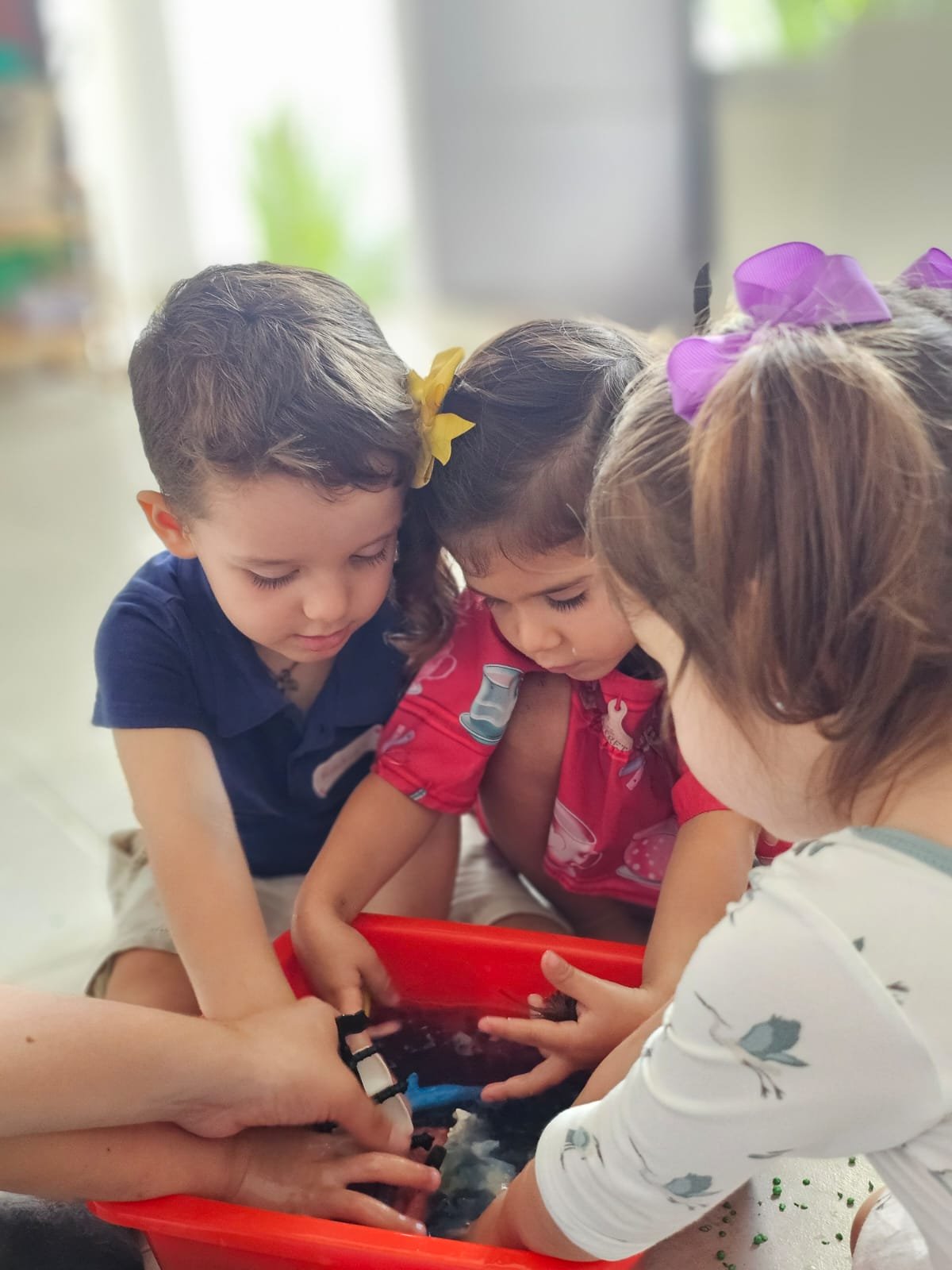 Niños jugando en un recipiente con agua, acompañados por una adulta, en un ambiente interior.