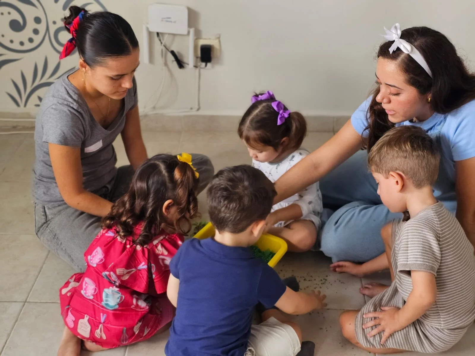 Tres mujeres y cinco niños sentados y acurrucados en el suelo, jugando con arena o un material similar en un ambiente interior.