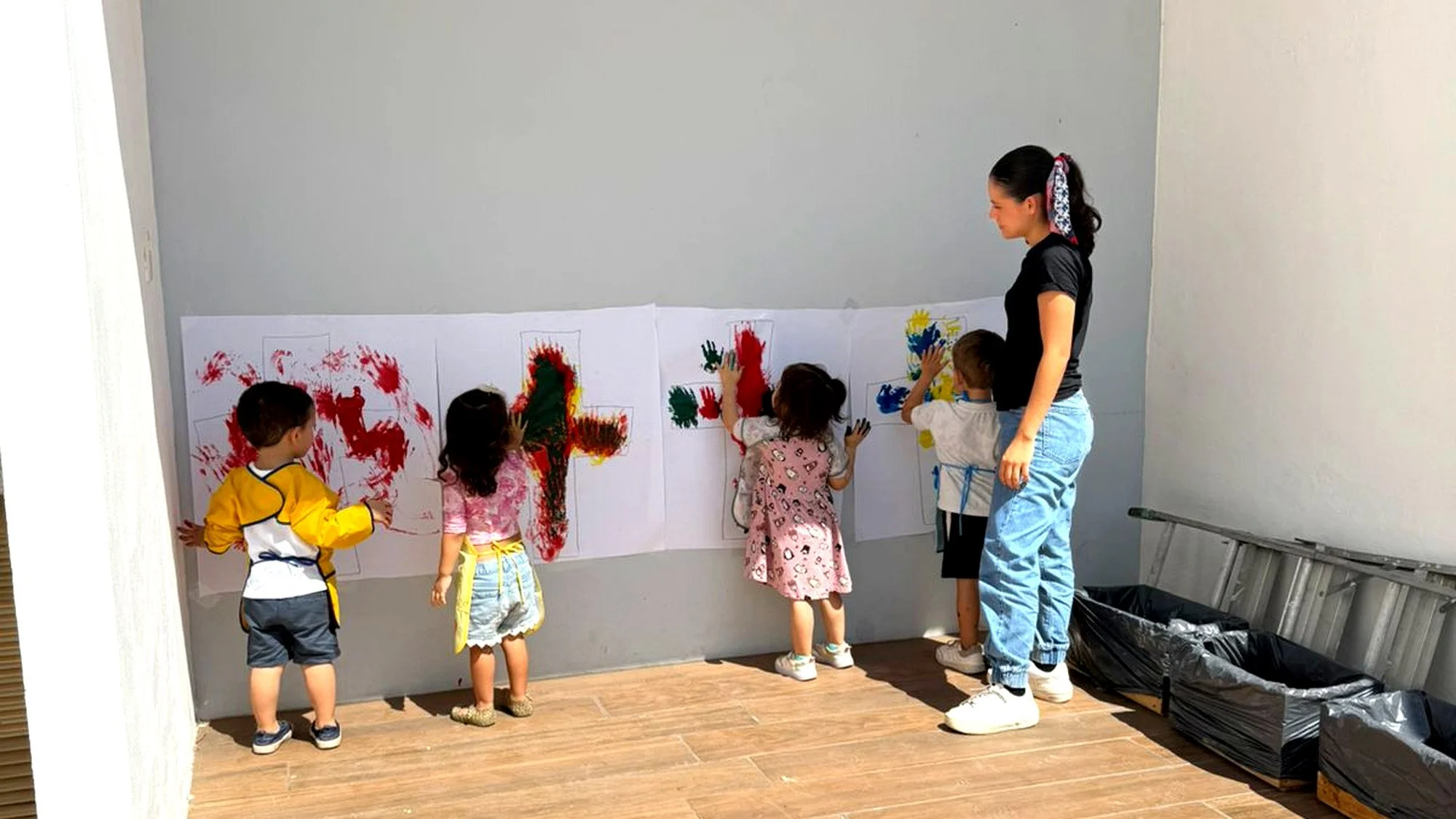Niños pintando en papel con tubos de pintura de colores, acompañados por una adulta en un ambiente interior.
