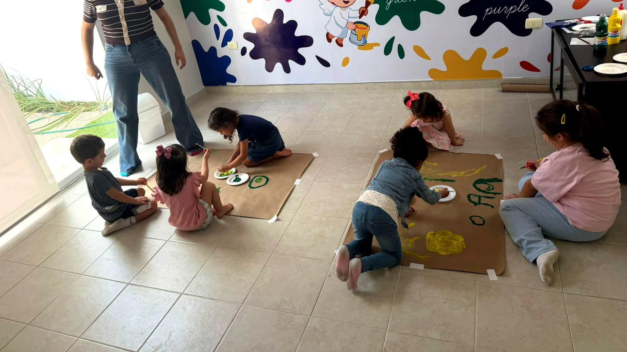 Niños y niñas pintando en papel en una clase de arte, con un mural colorido en la pared y una mesa con pinturas al fondo.