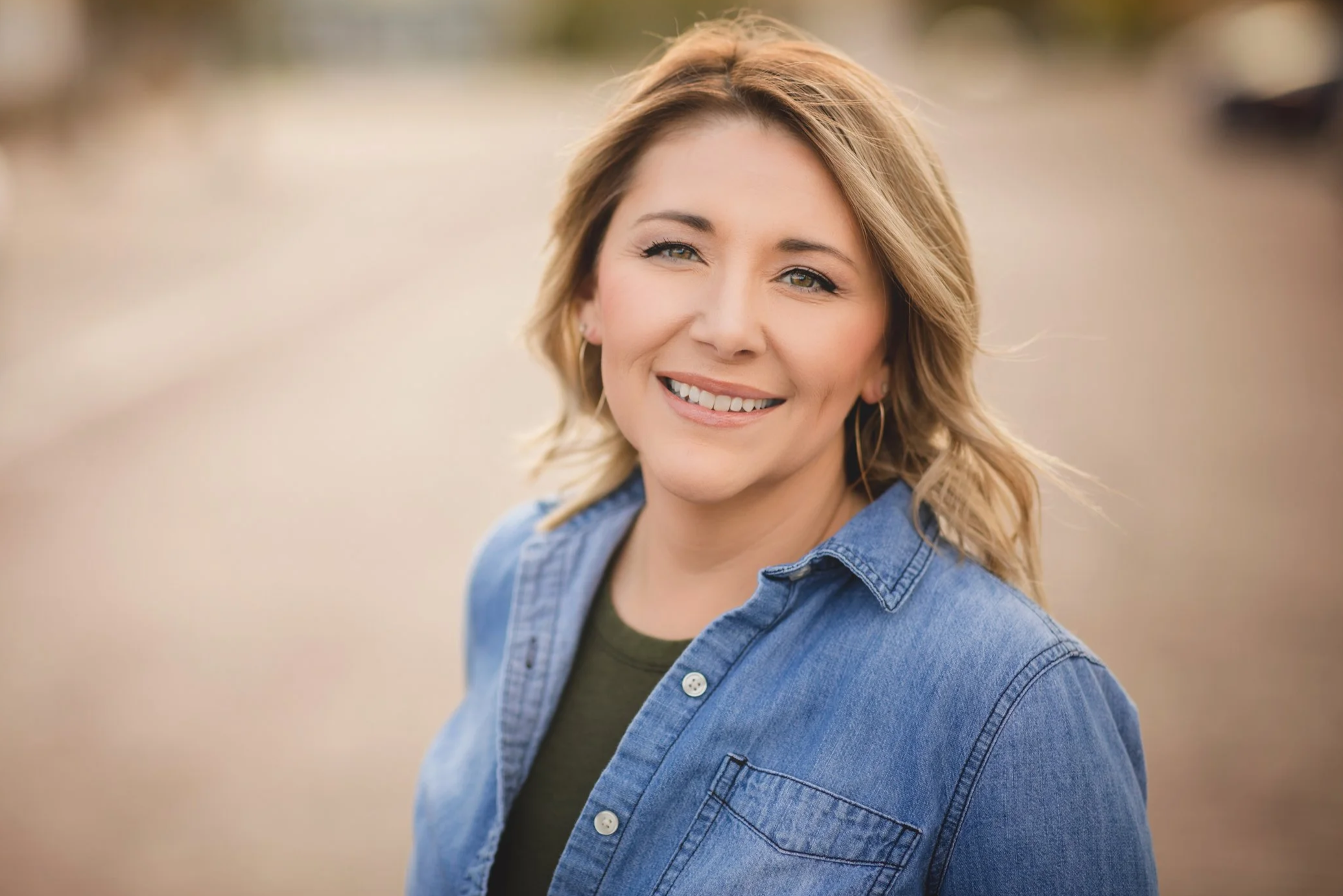 A woman with blonde hair smiling, wearing a blue denim shirt and hoop earrings, outdoors during daytime.