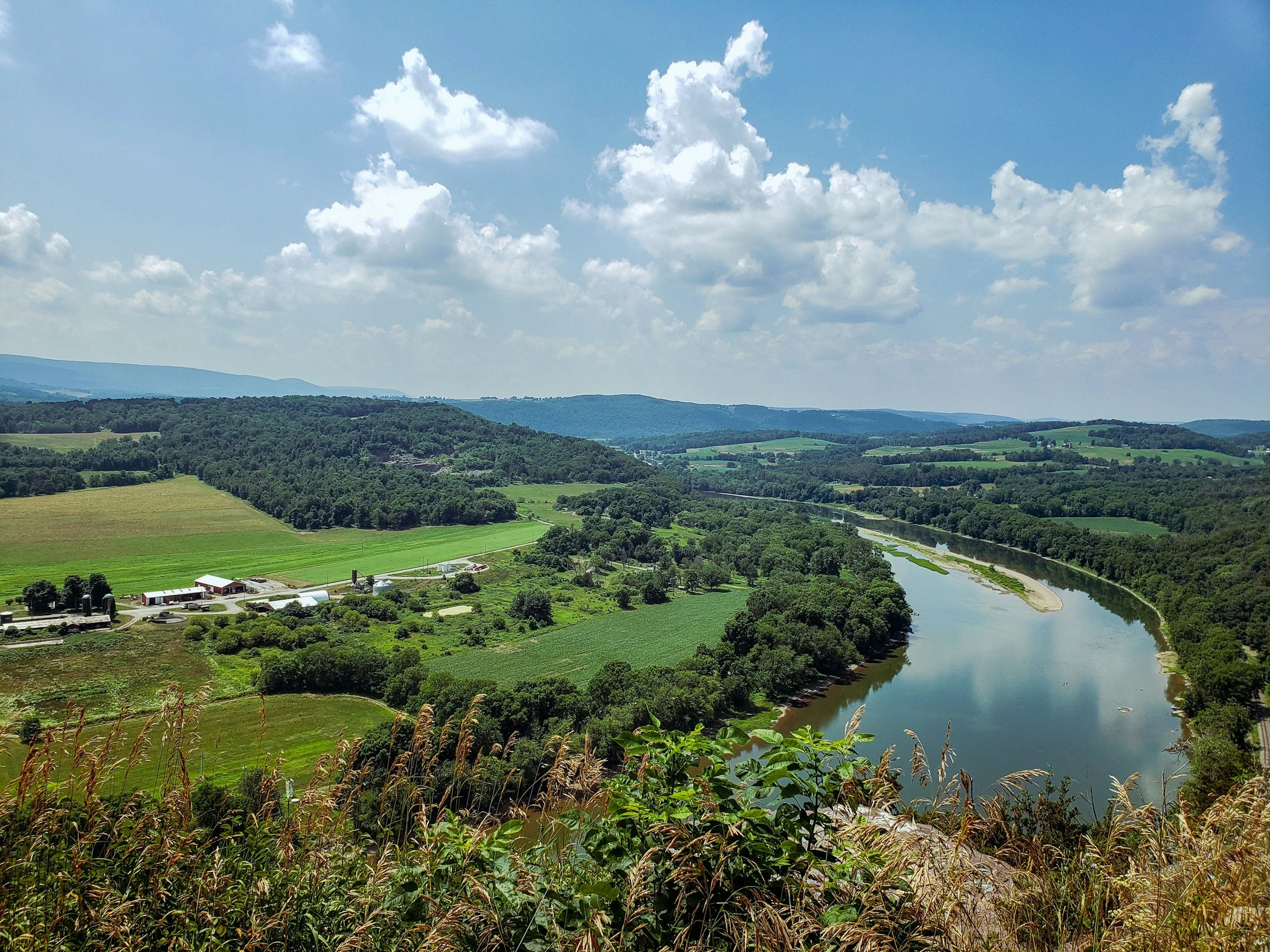 A scenic landscape with a winding river flowing through green fields, trees, and hills under a partly cloudy sky.