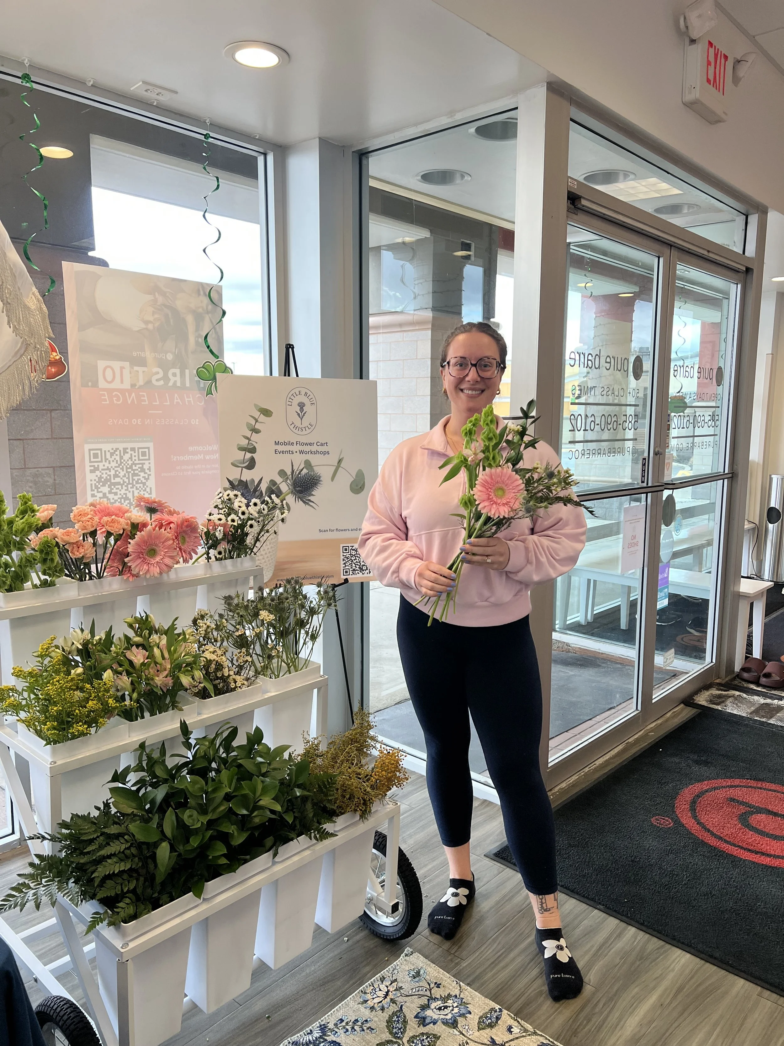 A woman standing inside a flower shop holding pink flowers, smiling, with various flowers displayed on white racks beside her.