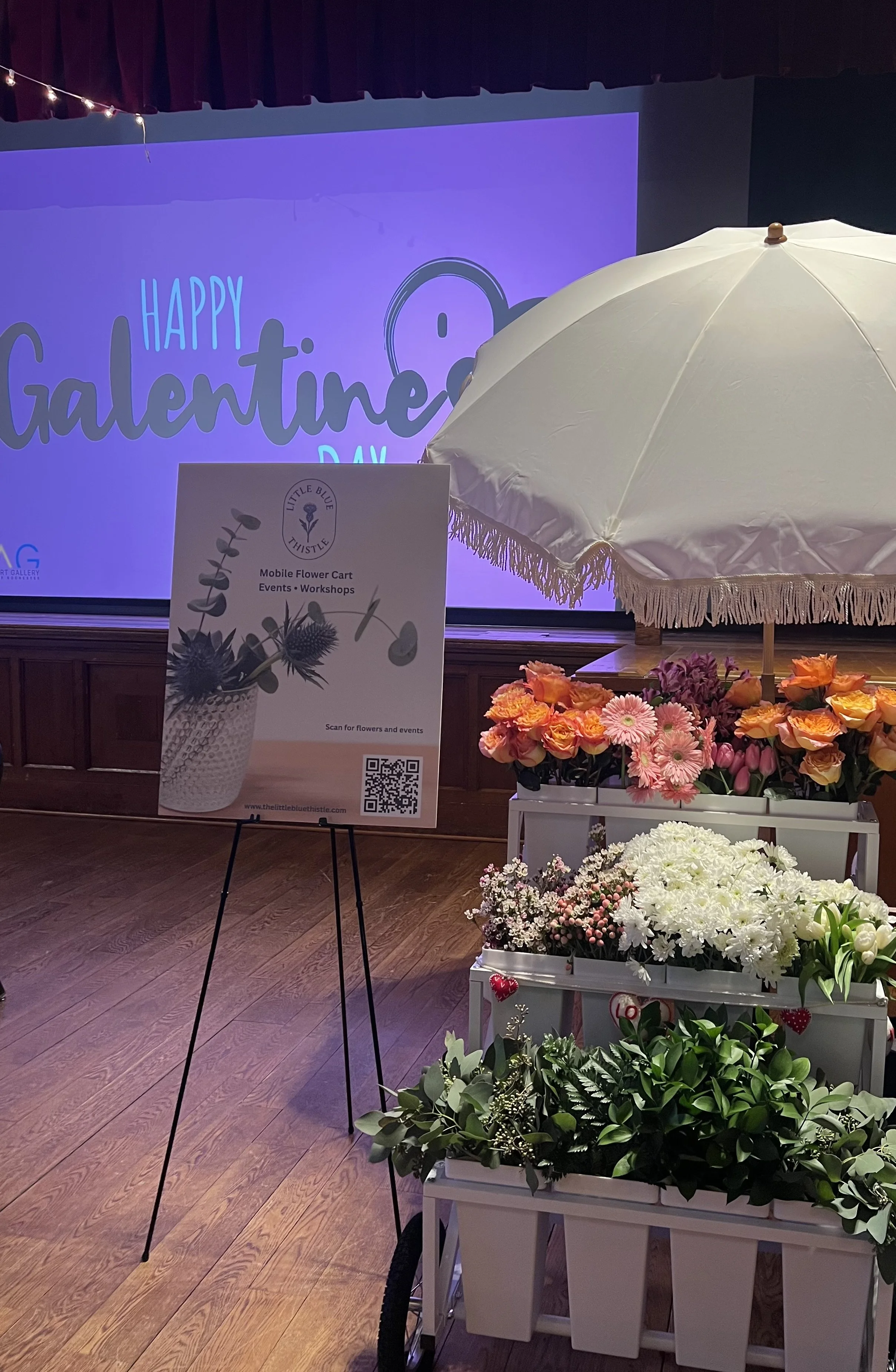 A flower cart with colorful pink, orange, white, and purple flowers under a white umbrella, set against a background with a stage and a large screen that says 'Happy Galentines Day'.