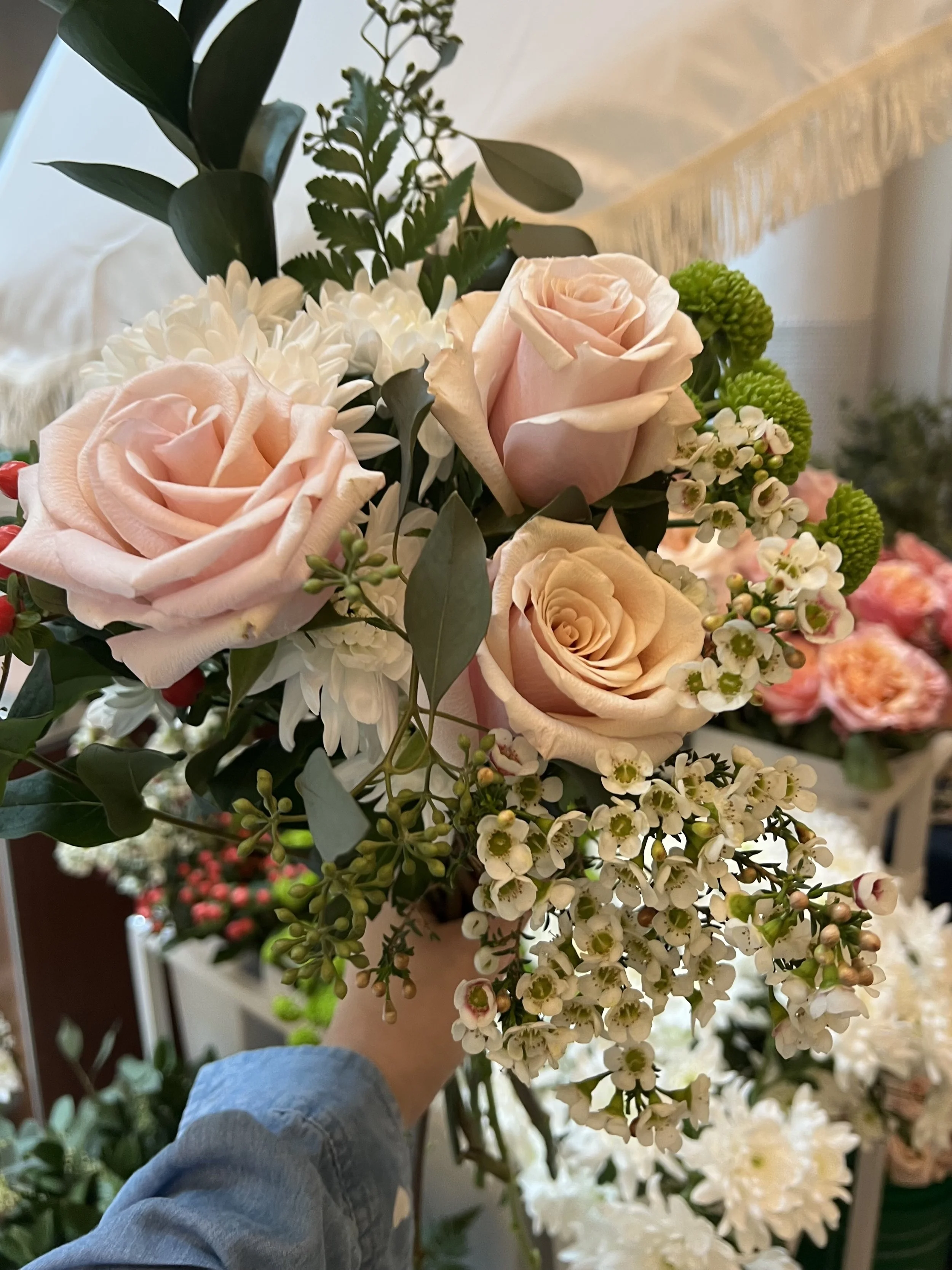 A person holding a bouquet of pink roses, white chrysanthemum, small white waxflowers, green button poms, and greenery.
