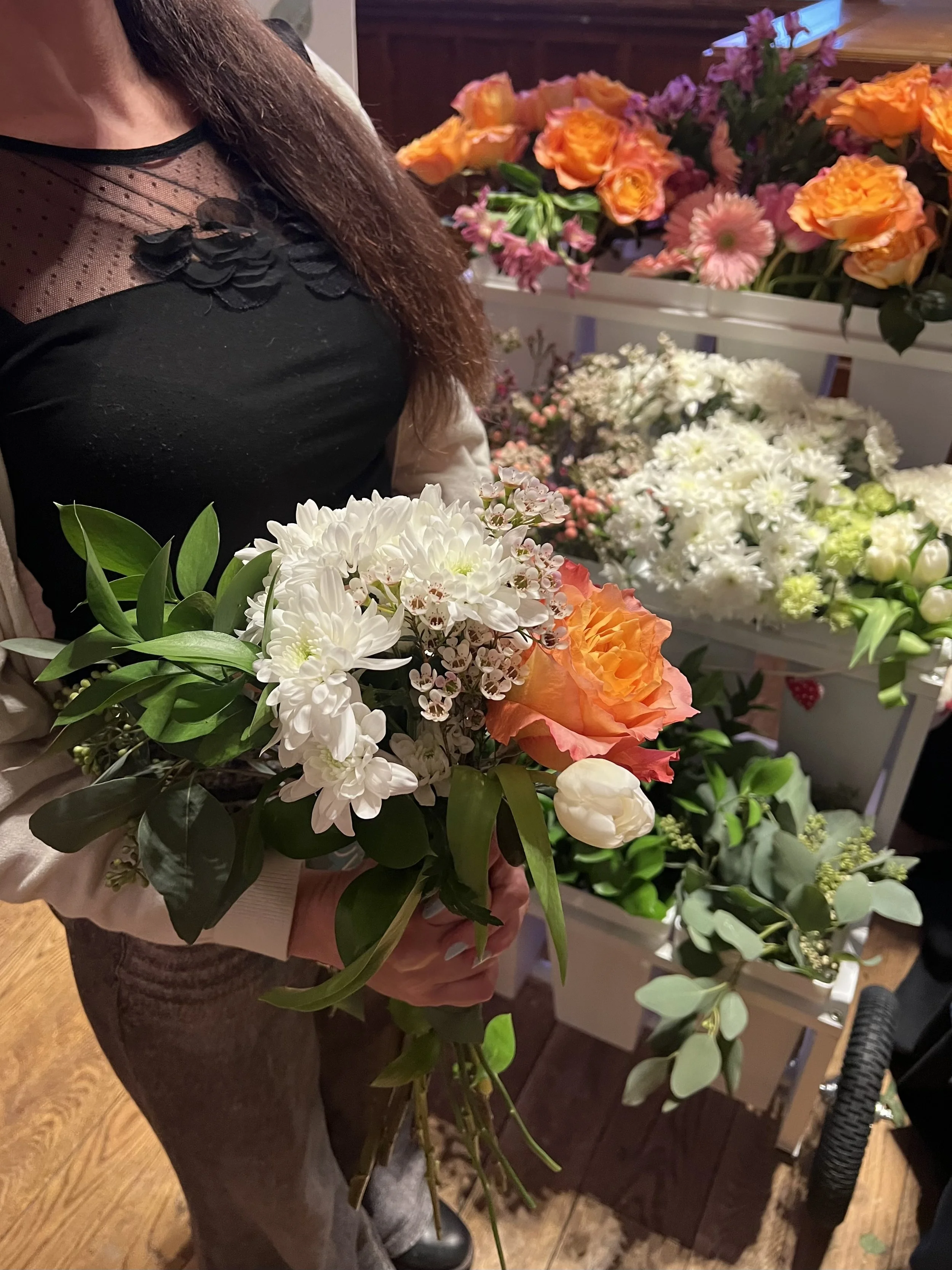 A woman holding a bouquet of various flowers, including white, orange, and pink blossoms, in a flower shop with other floral arrangements in the background.