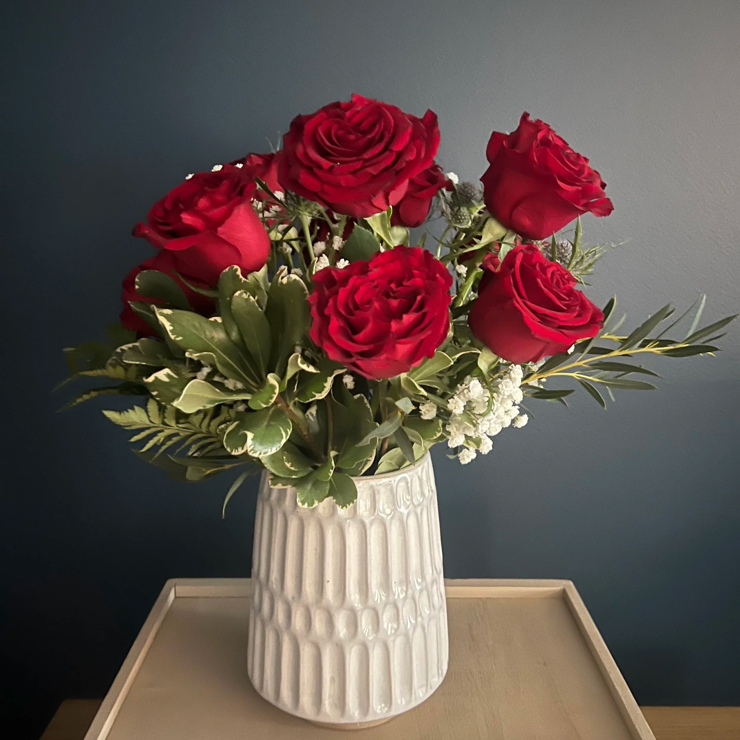 A bouquet of red roses, white filler flowers, and green foliage in a textured white ceramic vase on a light-colored table against a dark gray background.