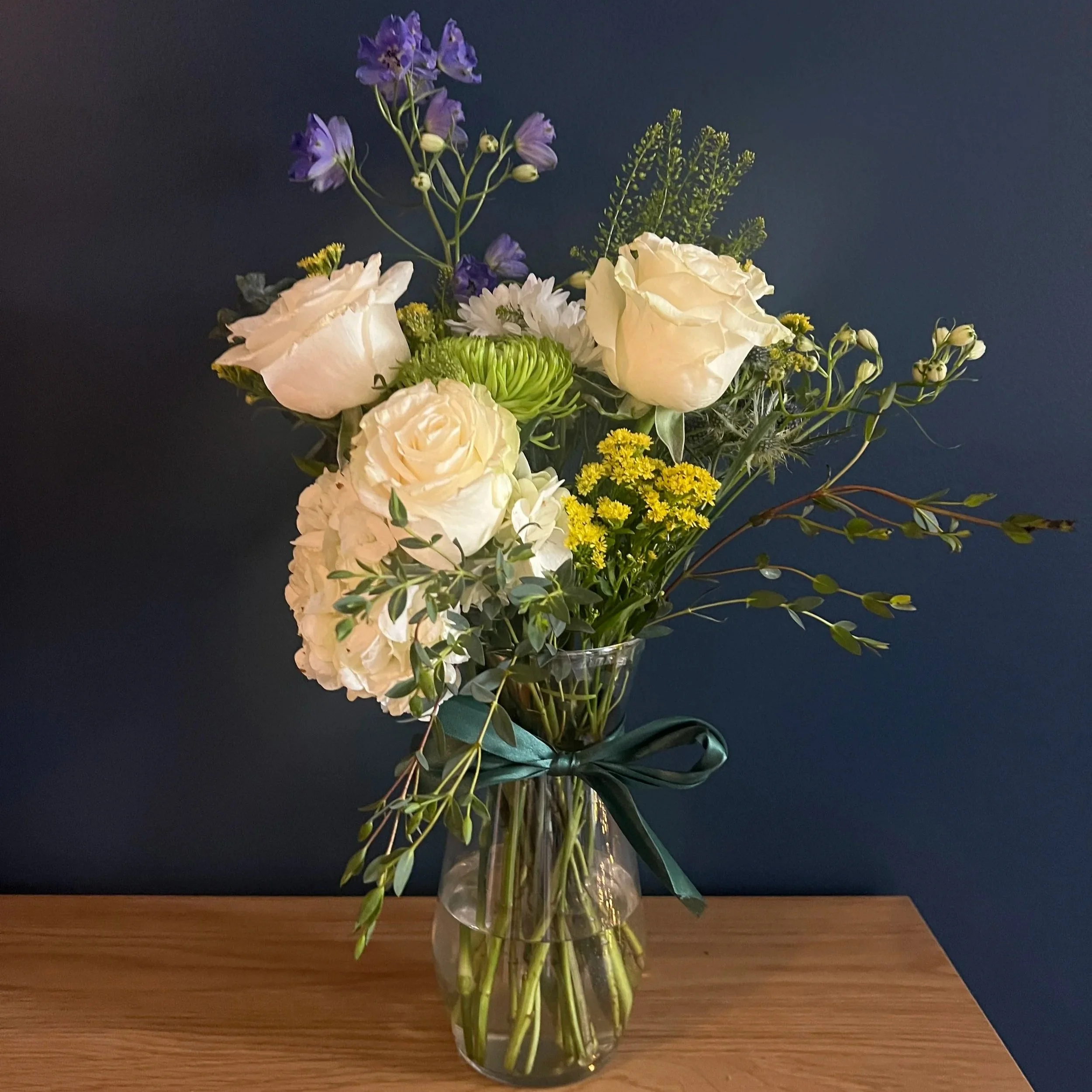 A floral arrangement in a glass vase with white roses, purple flowers, yellow blooms, greenery, and a teal ribbon, set on a wooden surface against a dark background.