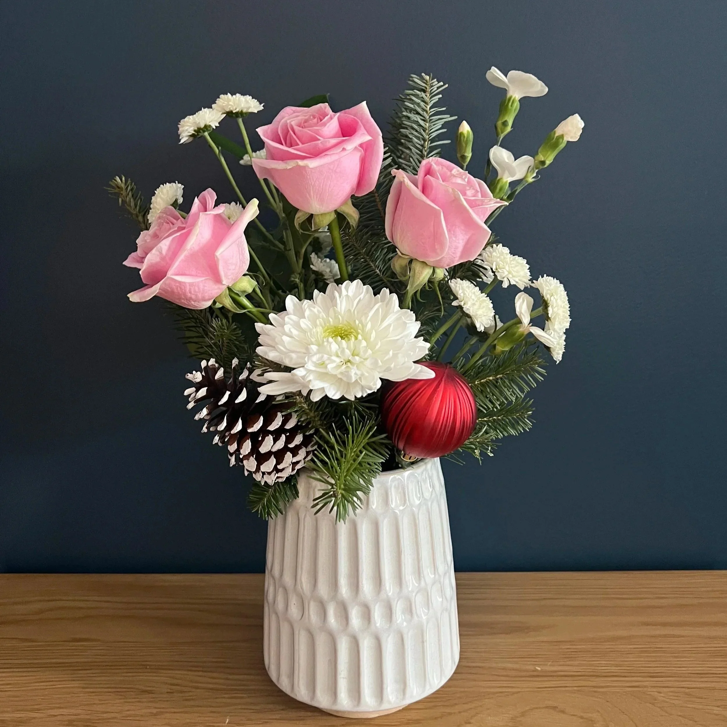 Flower arrangement with pink roses, white chrysanthemums, small white daisies, pinecones, and greenery in a white textured vase on a wooden surface against a dark background.