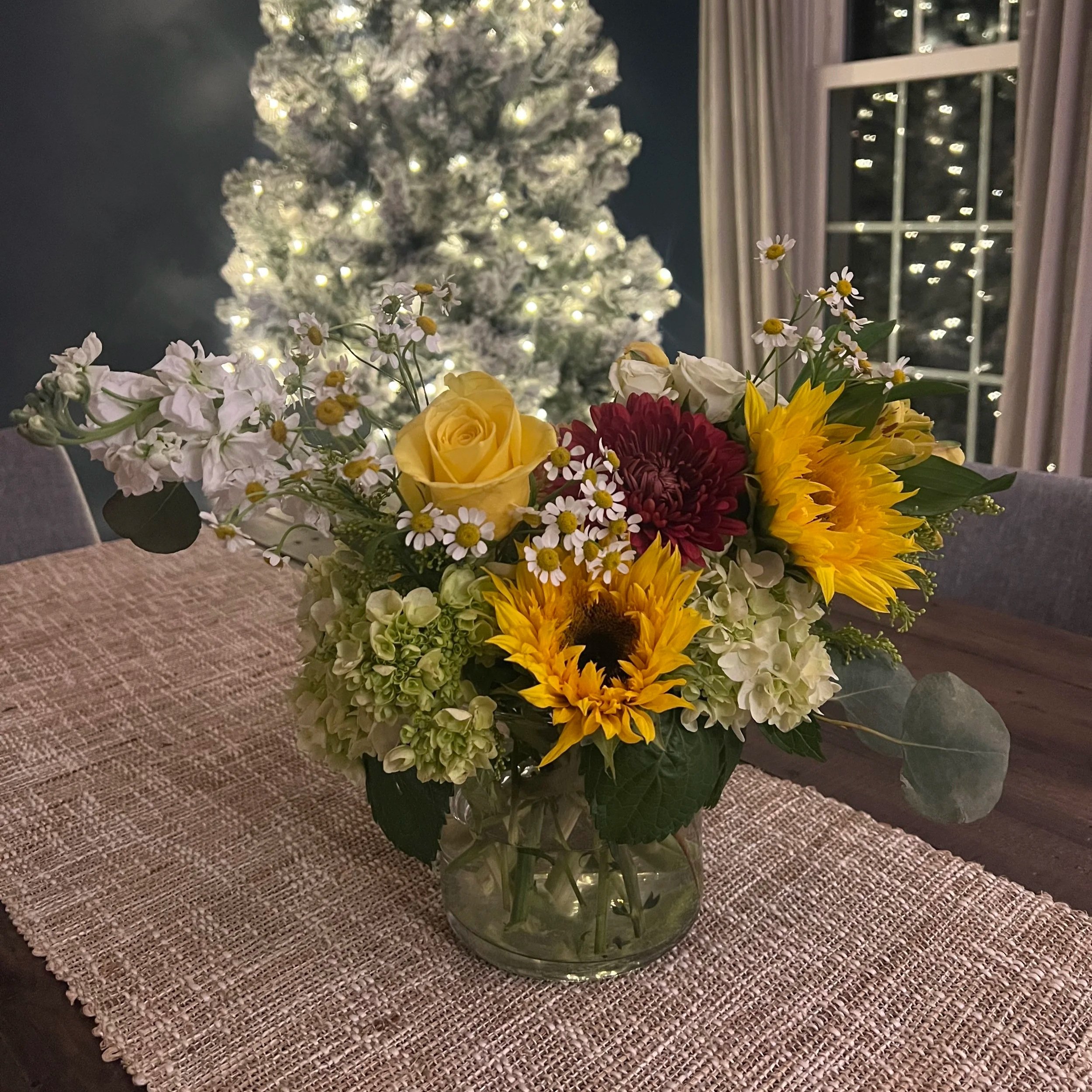 A colorful flower arrangement with yellow roses, sunflowers, red daisy, white daisy, and various greenery on a textured beige table runner in a dining room, with a lit Christmas tree and window in the background.