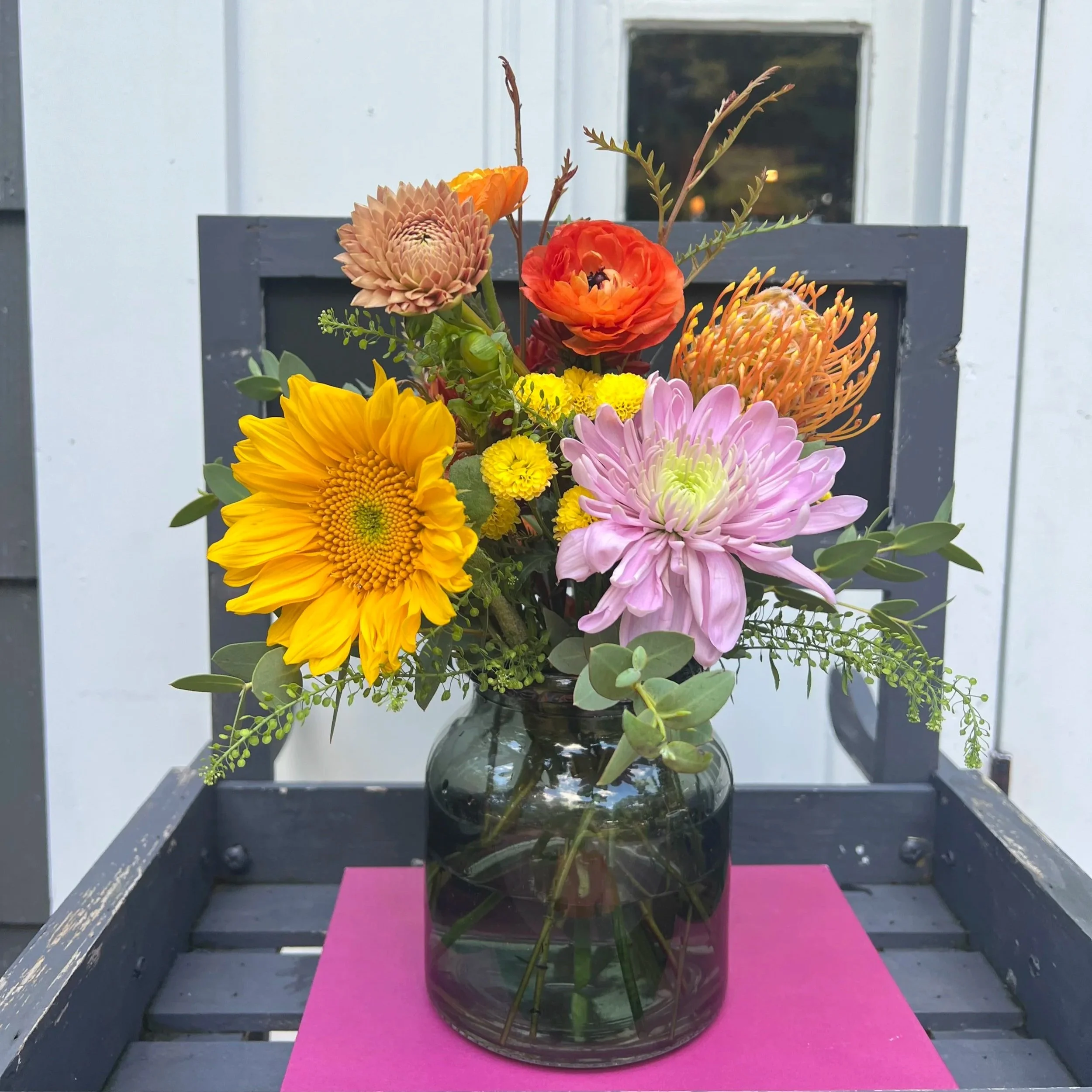 Colorful bouquet of various flowers in a glass vase on a pink surface outside, with a white fence in the background.