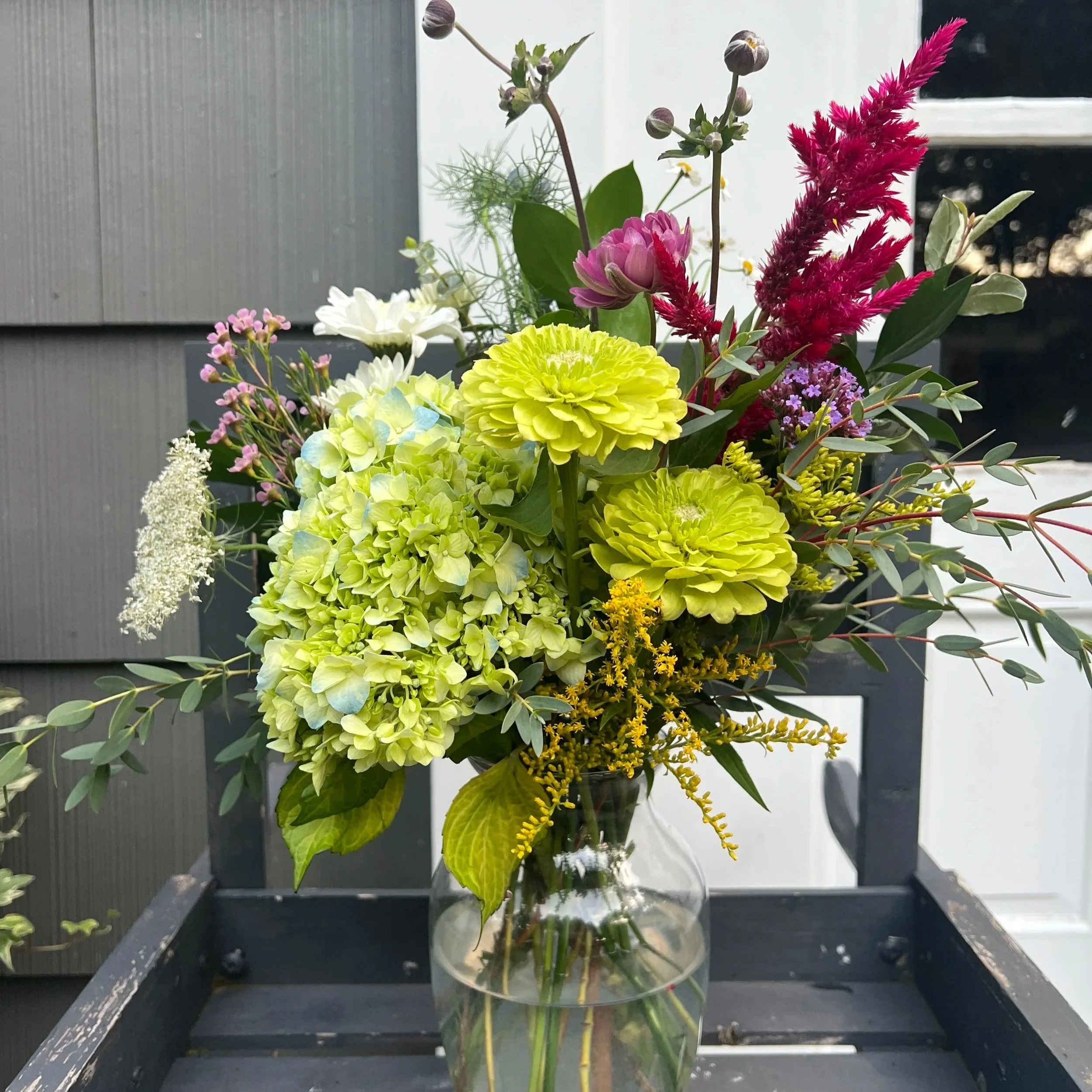 A colorful bouquet of mixed flowers in a glass vase on a black table outside, with gray panels and a white door in the background.