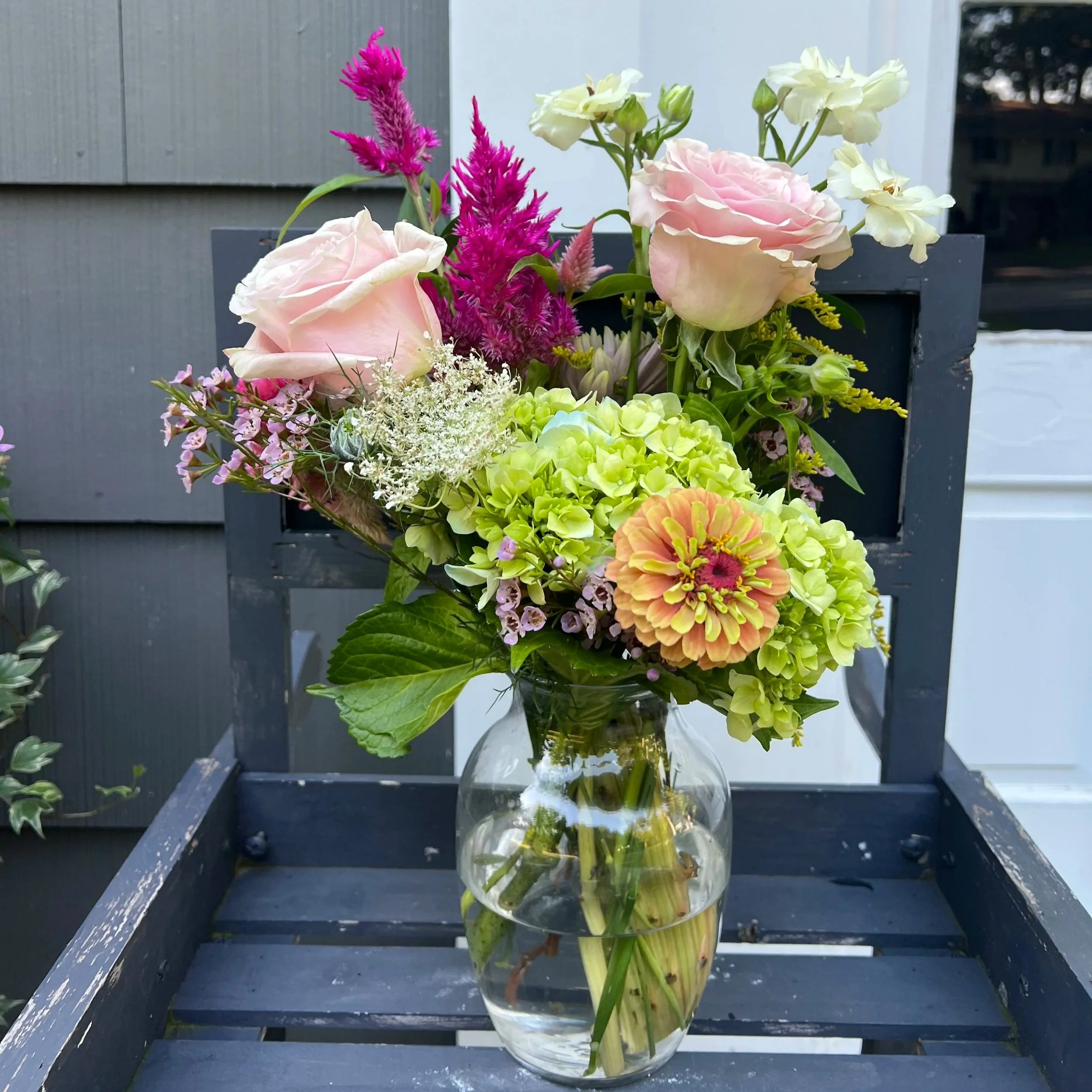 A bouquet of pink roses, white flowers, green hydrangeas, bright pink and orange zinnias in a glass vase on a dark blue wooden chair.