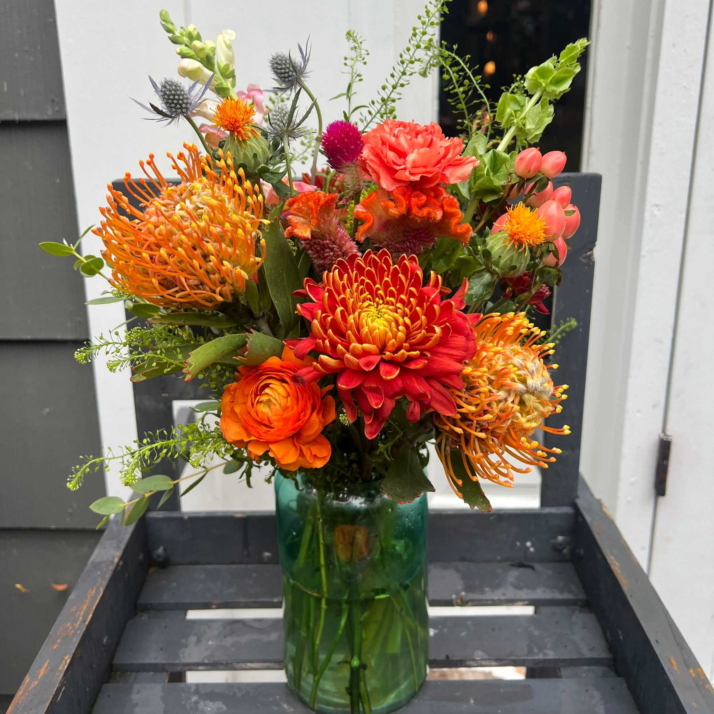 Vase of colorful mixed flowers including orange, red, and pink blooms, with greenery, on a wooden chair outdoors.