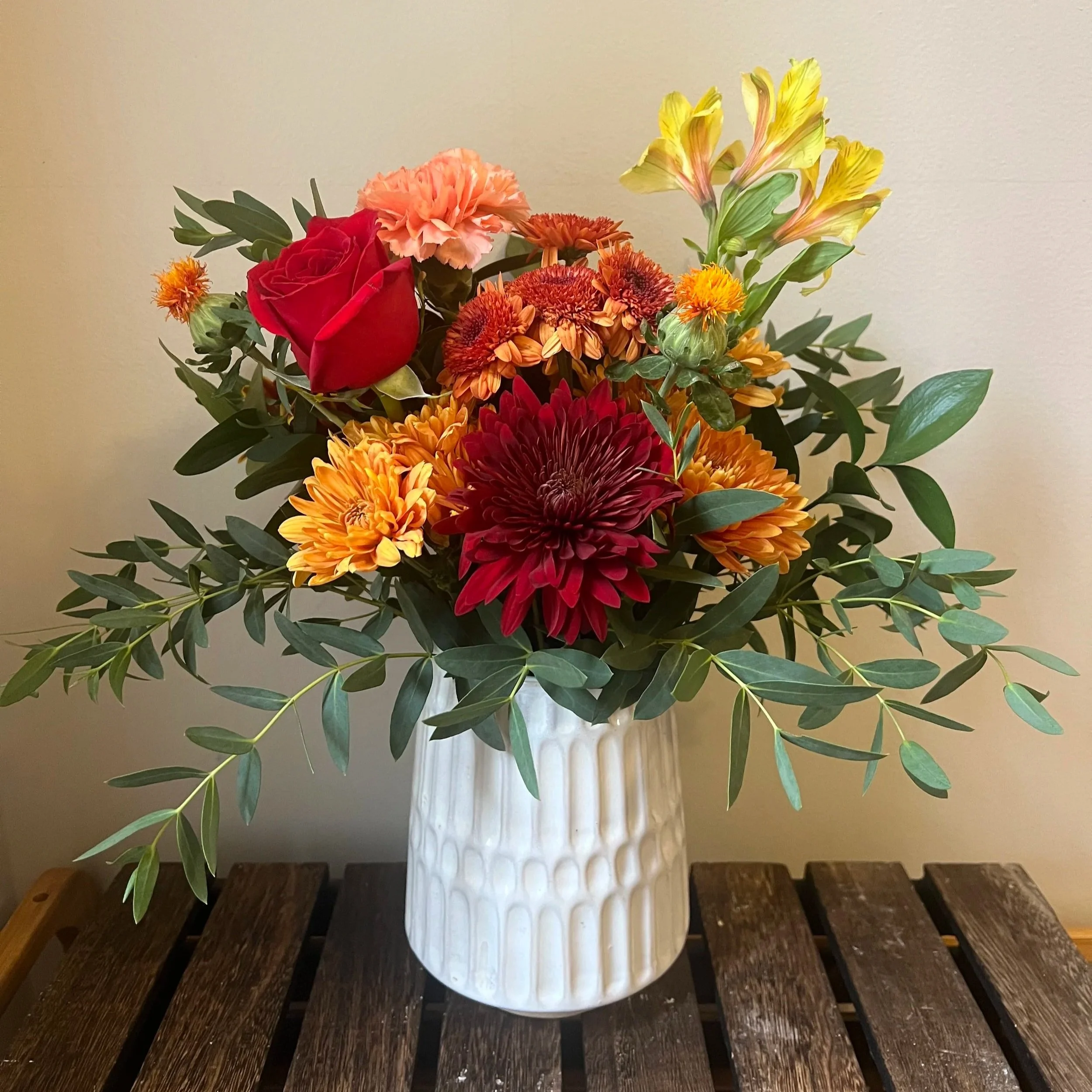 A bouquet of various colorful flowers in a white textured ceramic vase on a dark wooden surface against a plain beige wall.