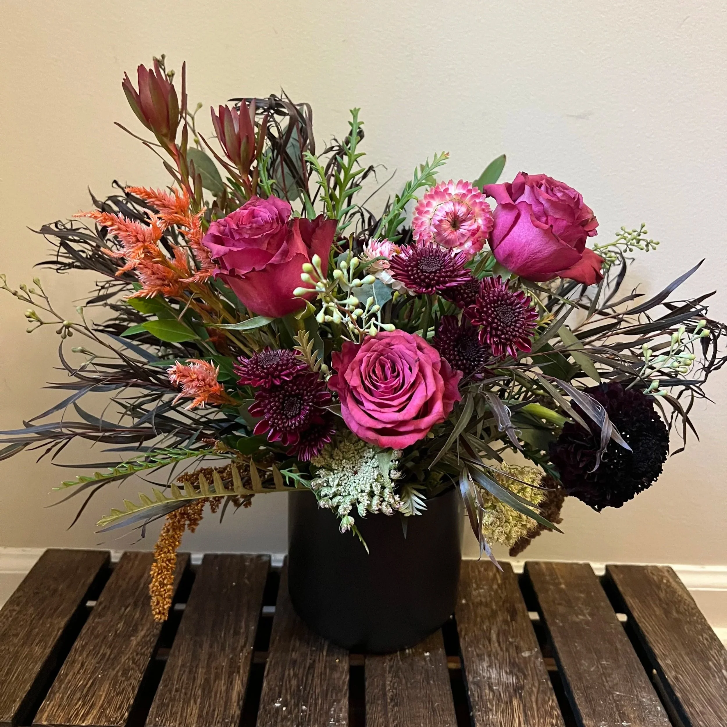 A floral arrangement in a black vase on a wooden surface with roses, chrysanthemums, and various greenery.