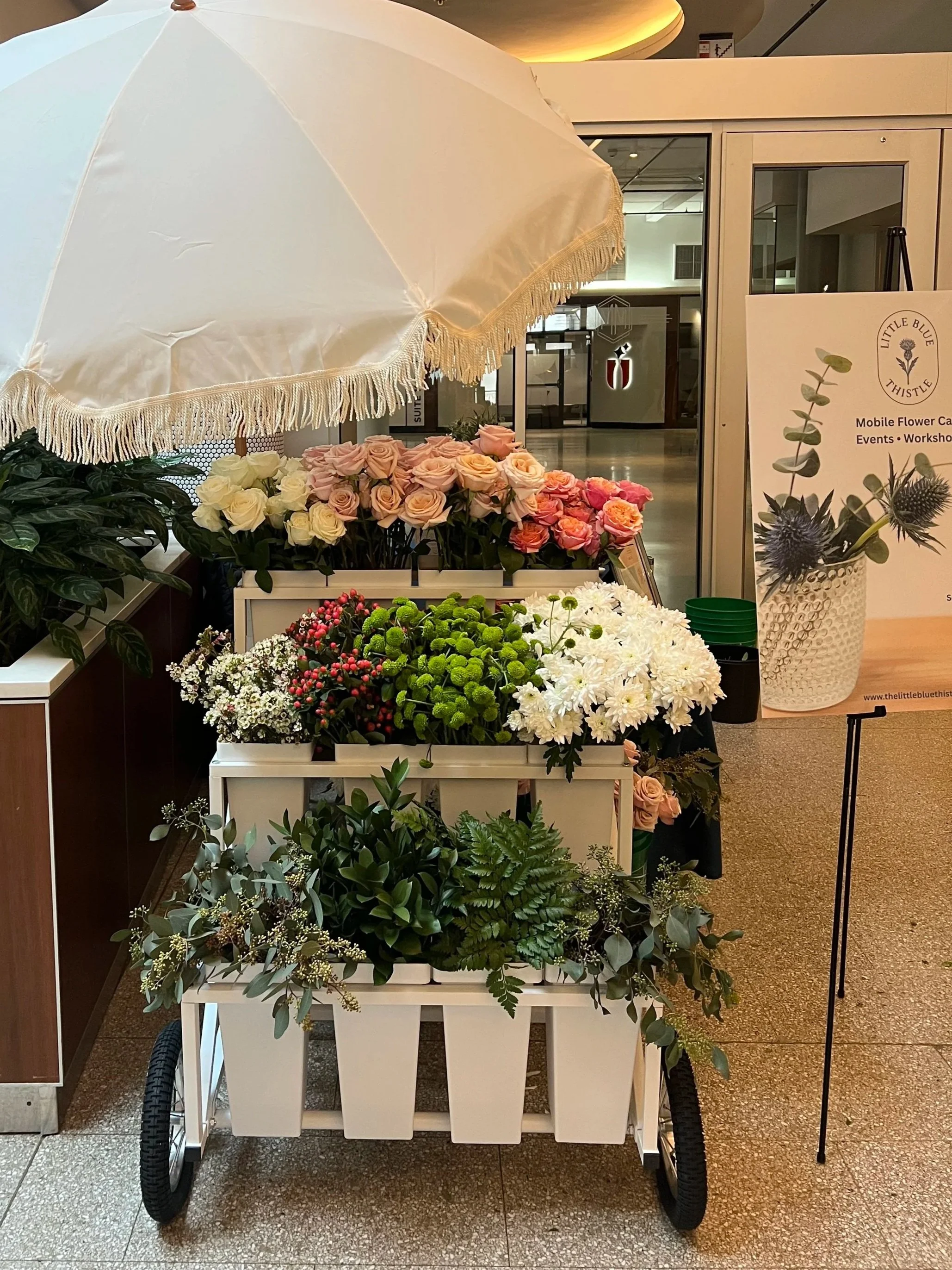 A white mobile flower cart with various flowers, including roses, chrysanthemums, and greenery, under a white parasol with fringe, located indoors near a sign and glass door.